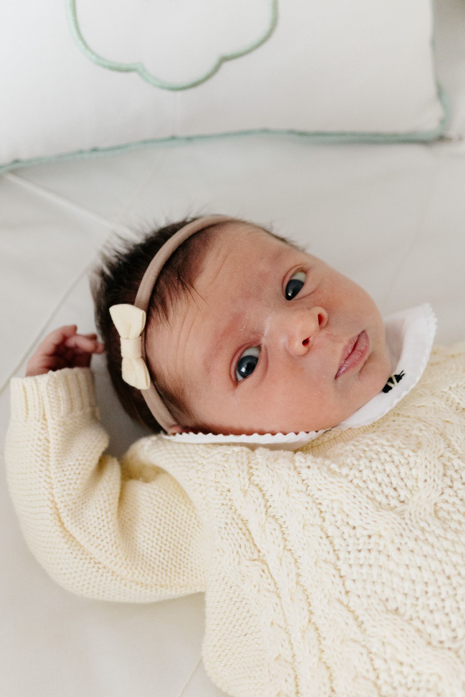 Newborn baby girl in cream knit sweater and bow headband lying on white bedding, looking at camera.