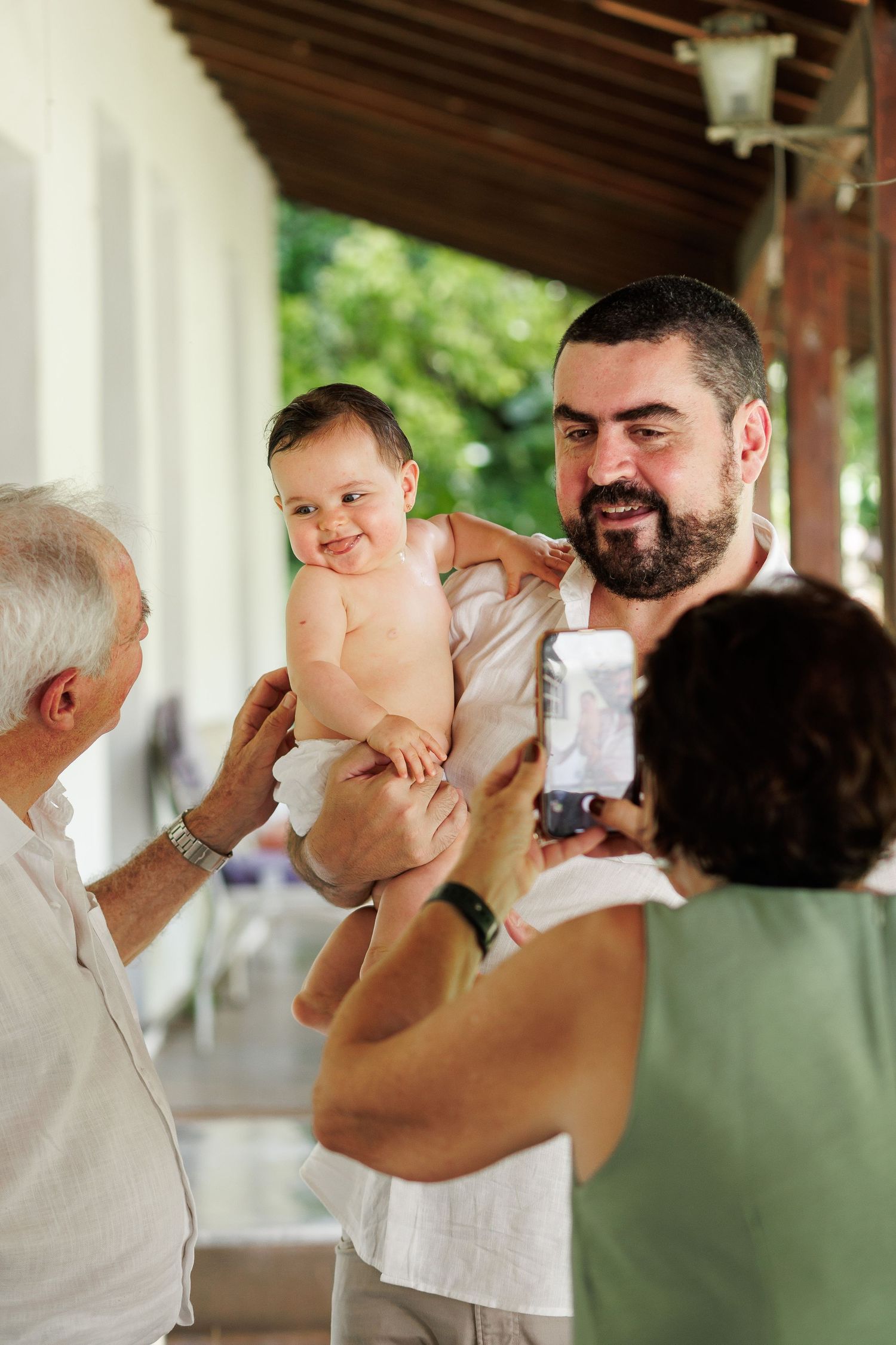 Bearded father holds smiling baby while elderly man and woman with phone gather on a sunny porch.