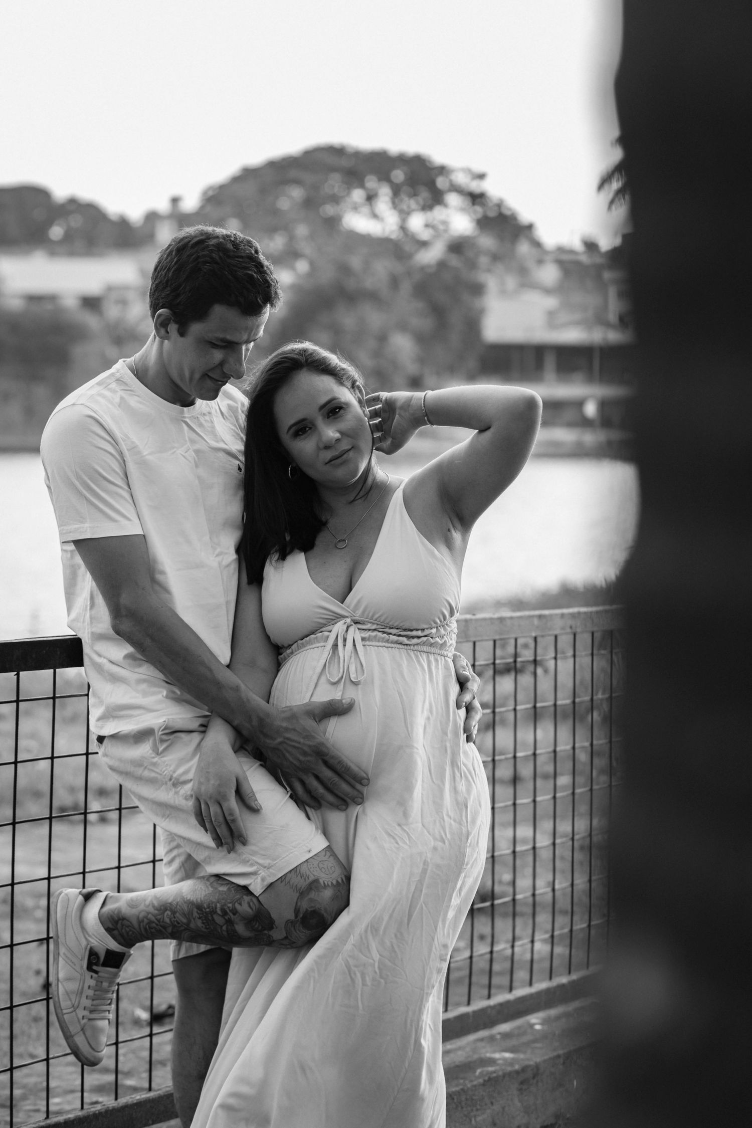 Black and white maternity portrait of couple posing by waterfront railing.