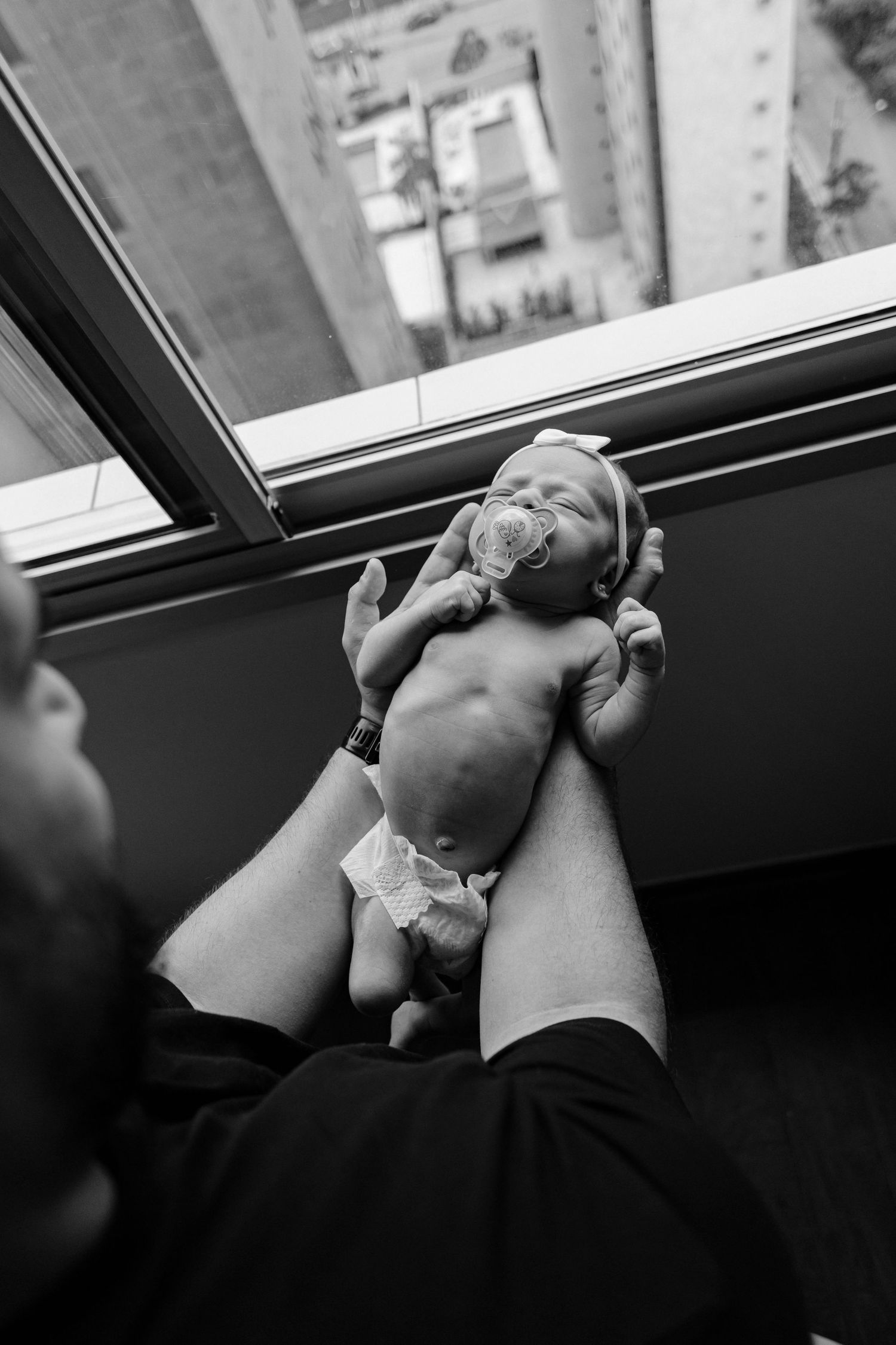 Black and white image of tiny newborn being held up against a window view.