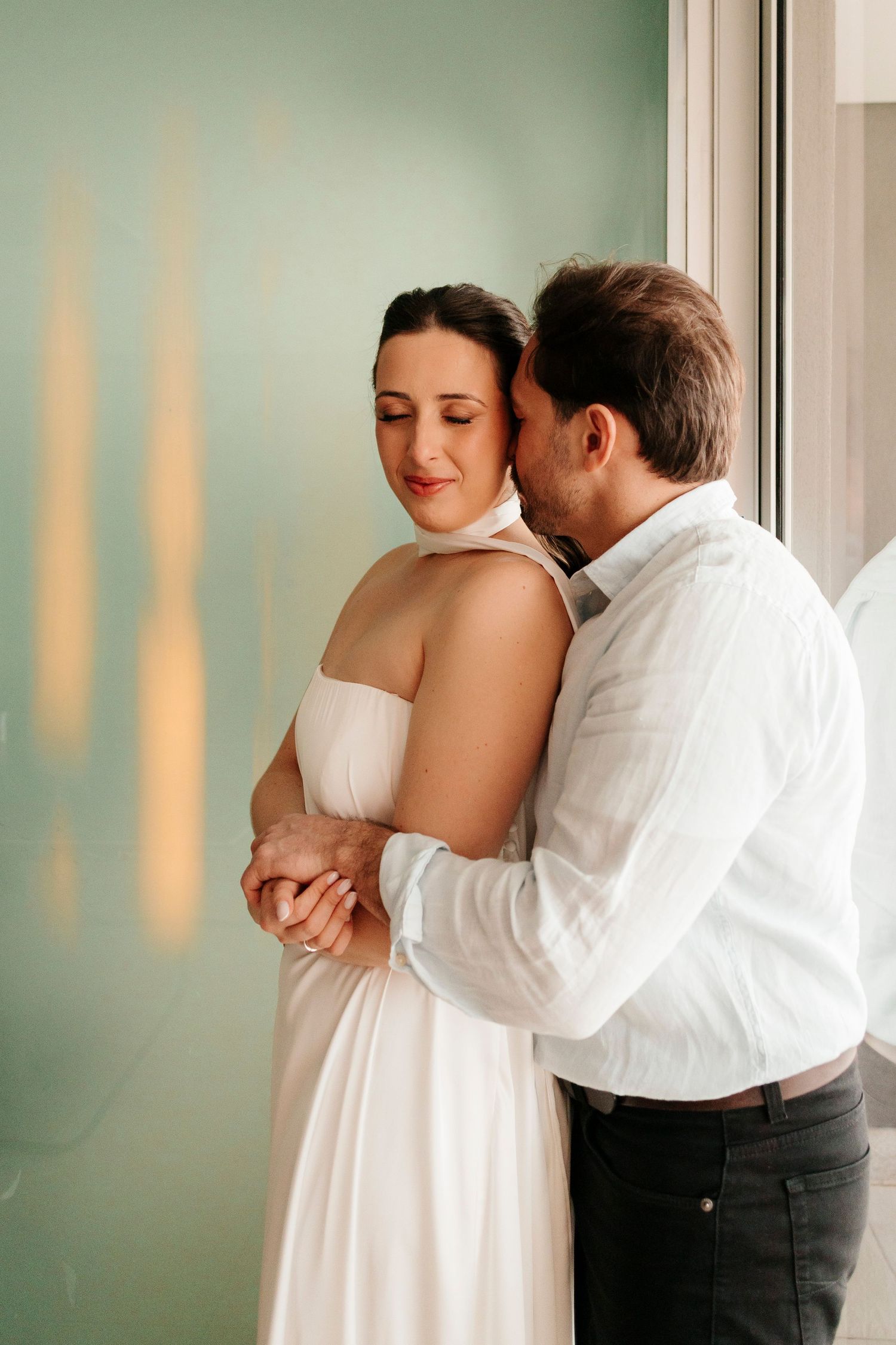 Intimate indoor portrait of couple in white and formal attire by window.