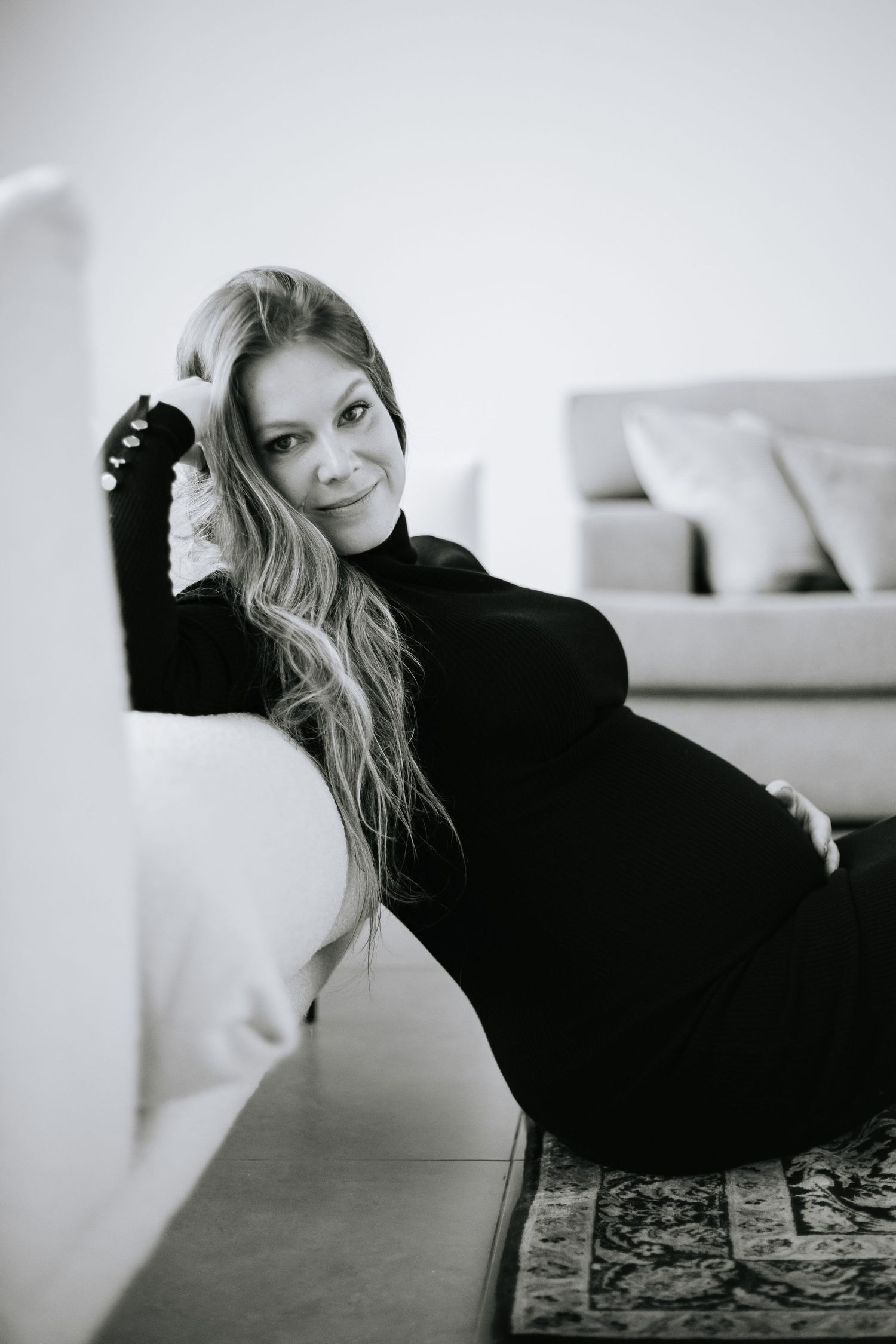 Monochrome portrait of expectant mother in black dress posing beside white sofa.