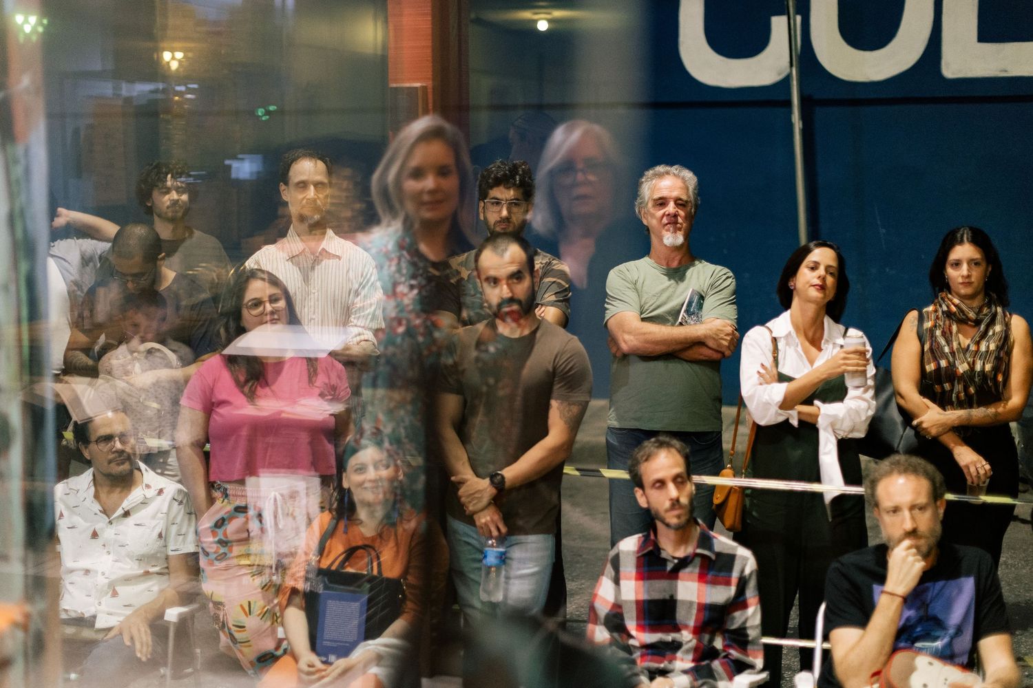 A diverse group of people watching an event through a glass window, reflecting urban surroundings in a city setting.
