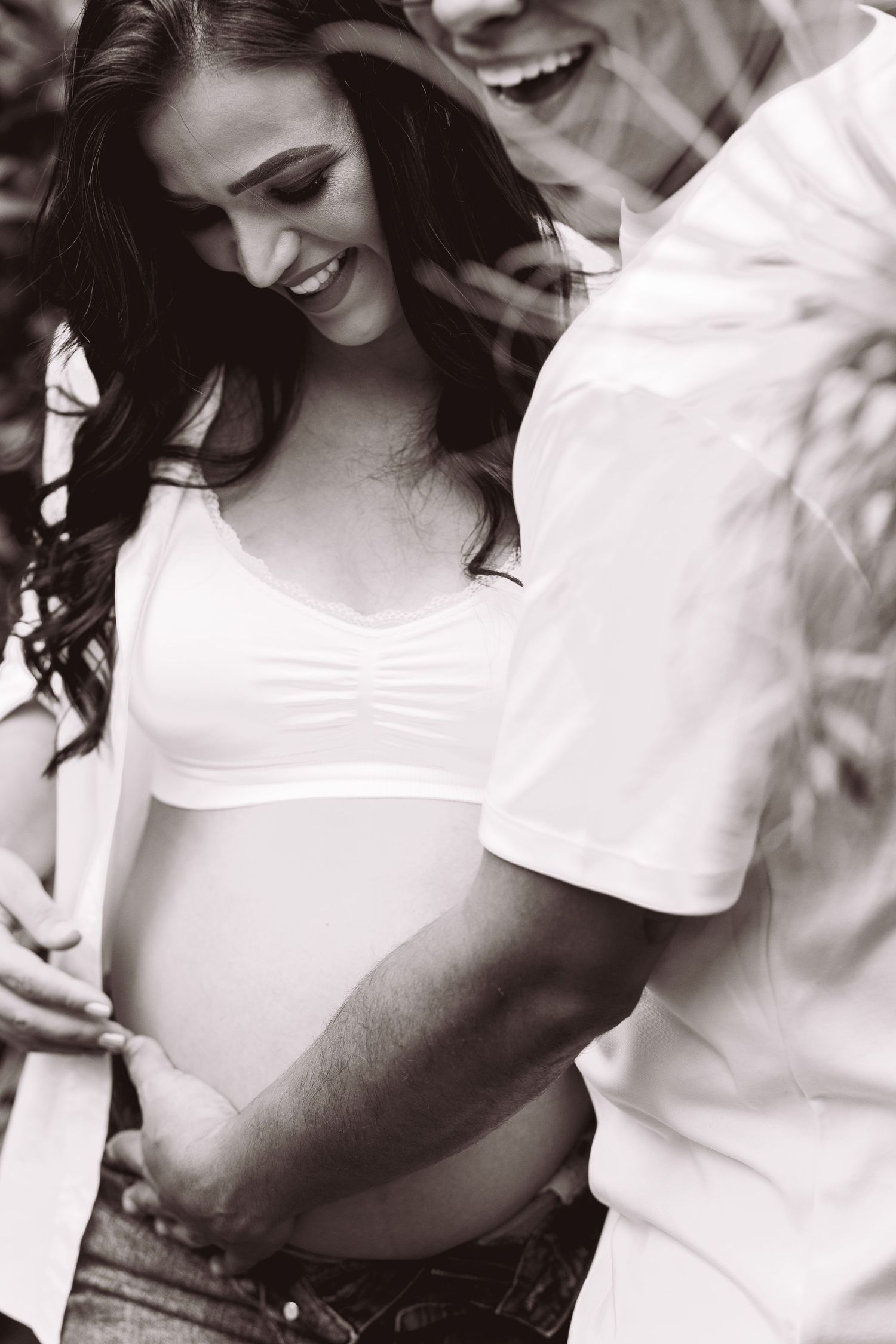 Black and white photo of a smiling pregnant woman and partner tenderly cradling her baby bump outdoors.