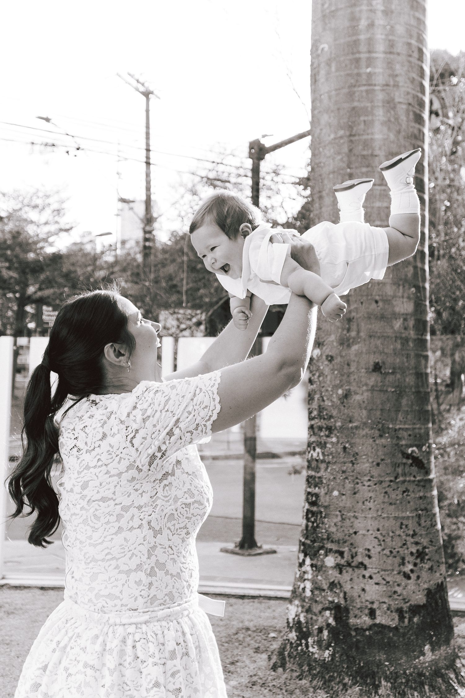 A woman in a white lace dress lifts a baby up in the air in front of a palm tree in a black and white photograph.