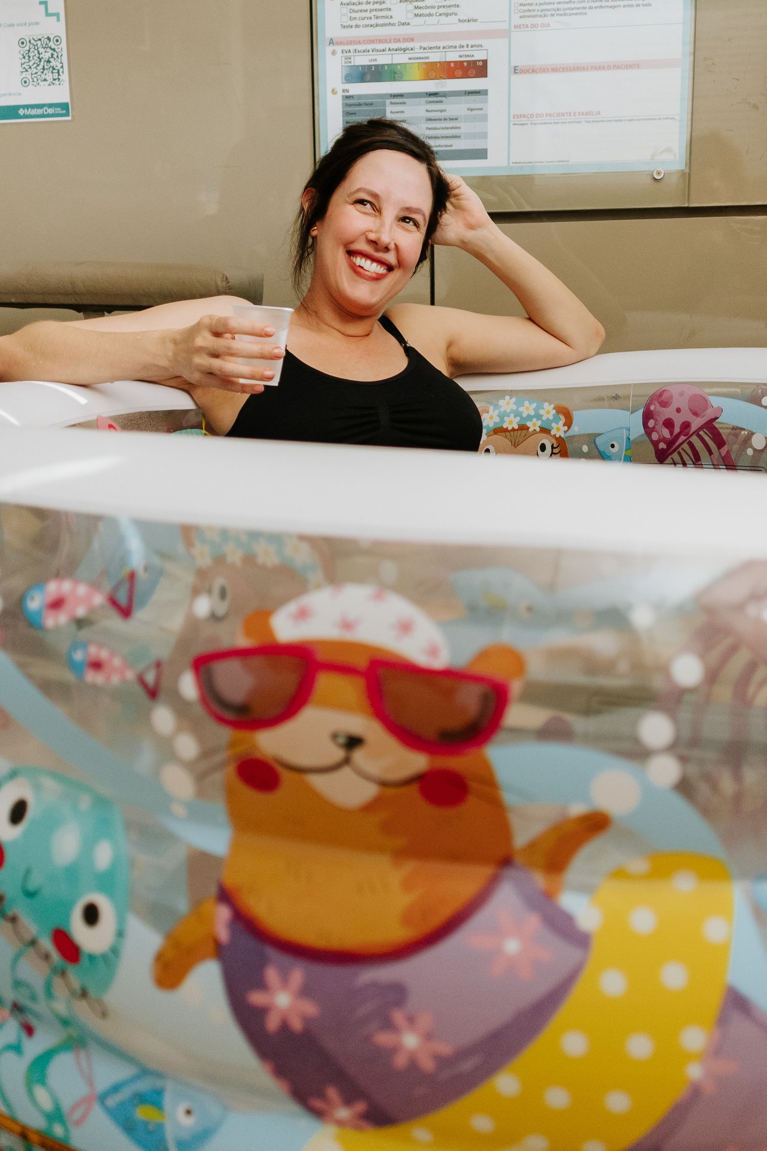Smiling woman in black swimsuit relaxing in a colorful inflatable pool with cartoon bear design, holding a drink.