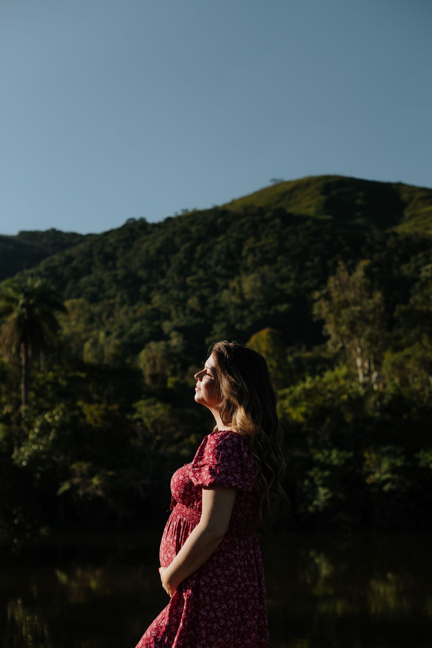 Pregnant woman in red dress poses against scenic mountain and lake backdrop.
