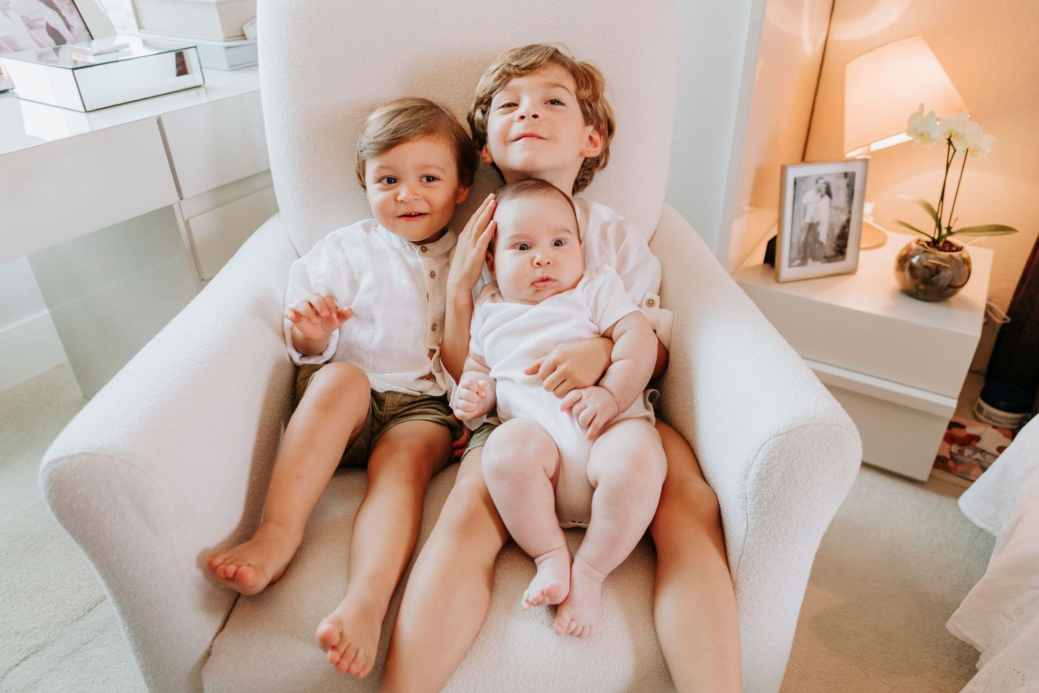 Three children relax together on a cozy white armchair in a warmly lit room with a lamp in the background.