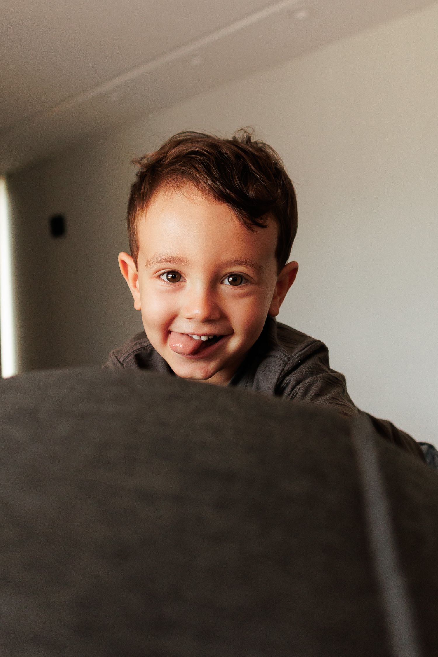 A cheerful young boy with dark hair peeks over a gray couch, smiling brightly in a well-lit room.