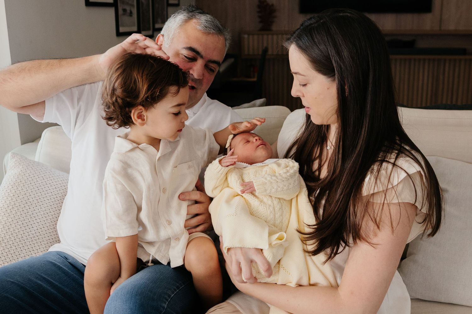 Family of four with newborn baby sharing a tender moment on a white sofa at home.