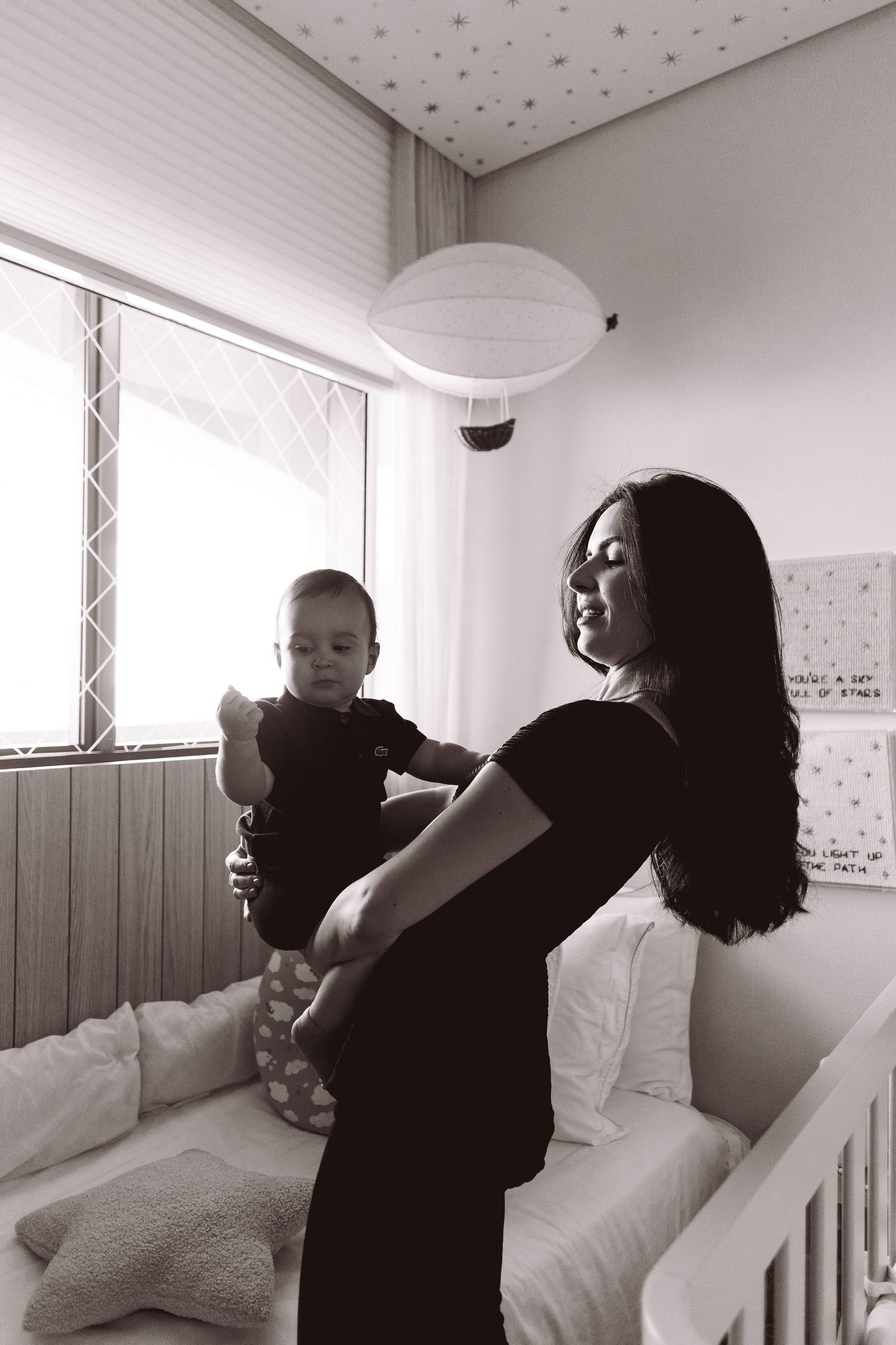 Black and white photo of a mother holding her baby in a nursery room with a crib and star-decorated ceiling.