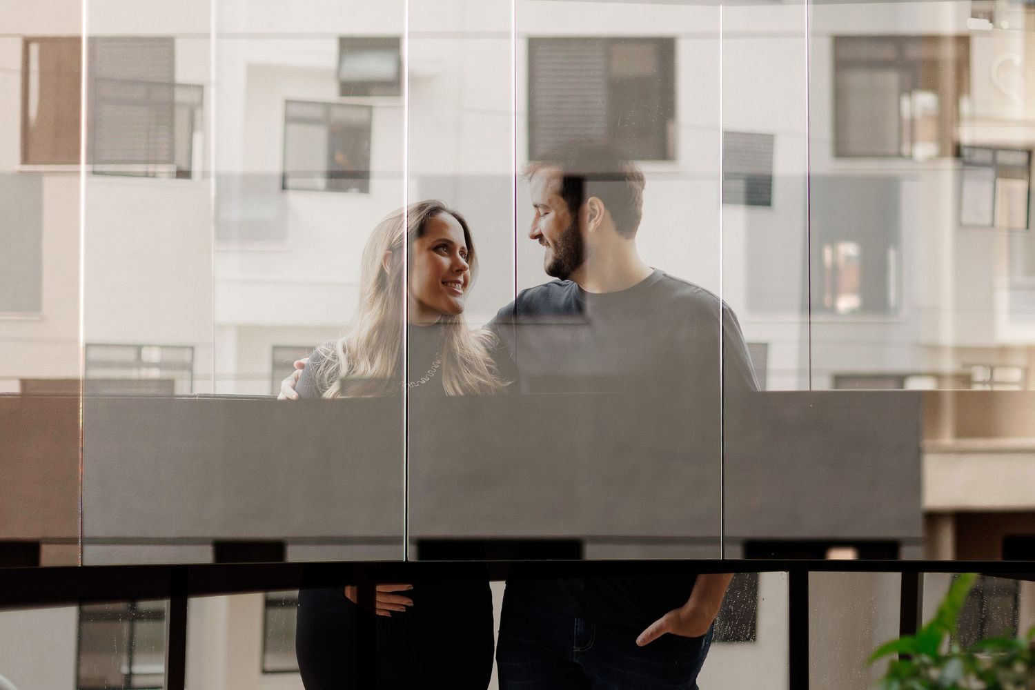 A smiling couple shares a romantic moment on a modern glass balcony with an urban apartment building in the background.