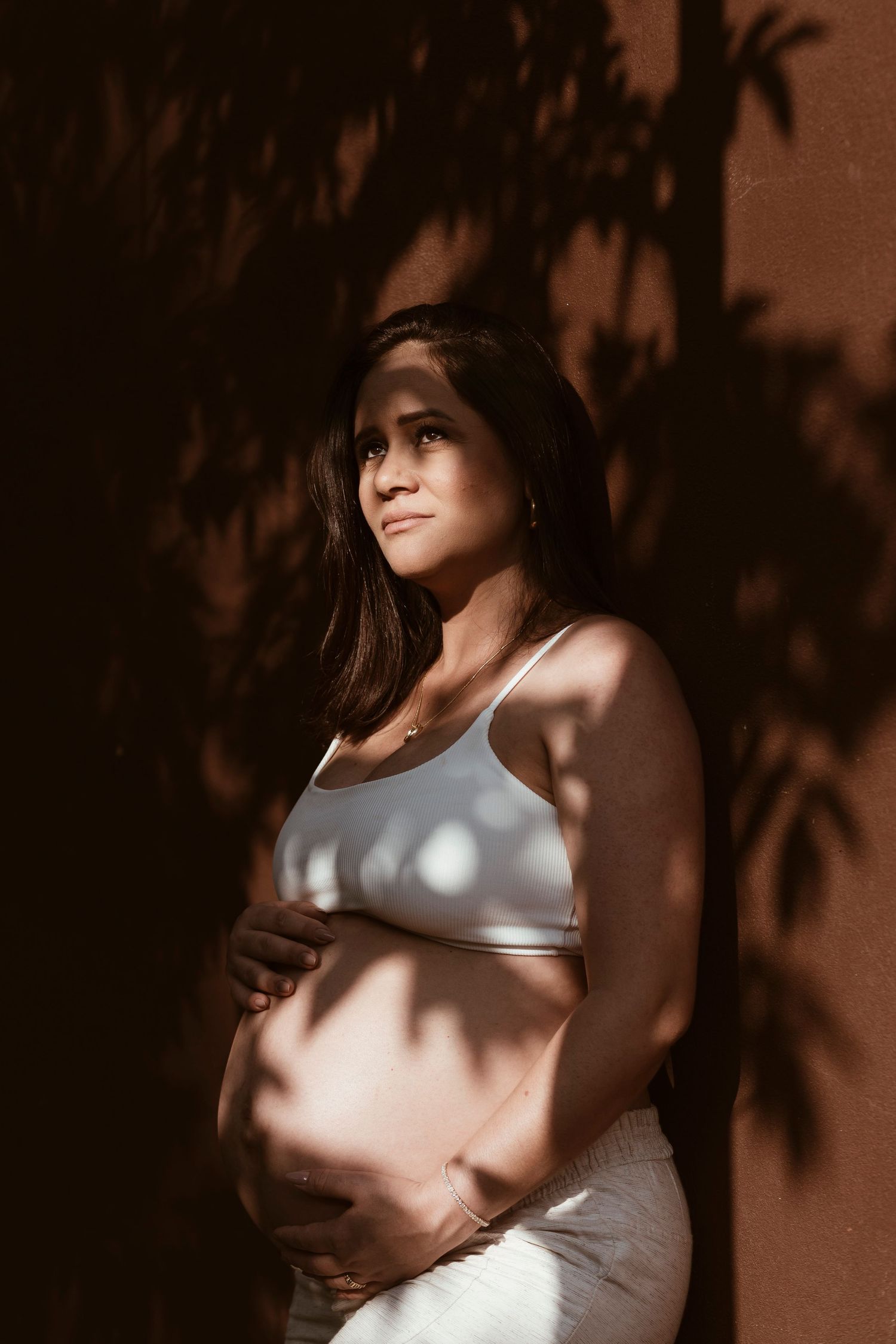 Artistic maternity portrait with palm tree shadows creating patterns on pregnant woman's silhouette.