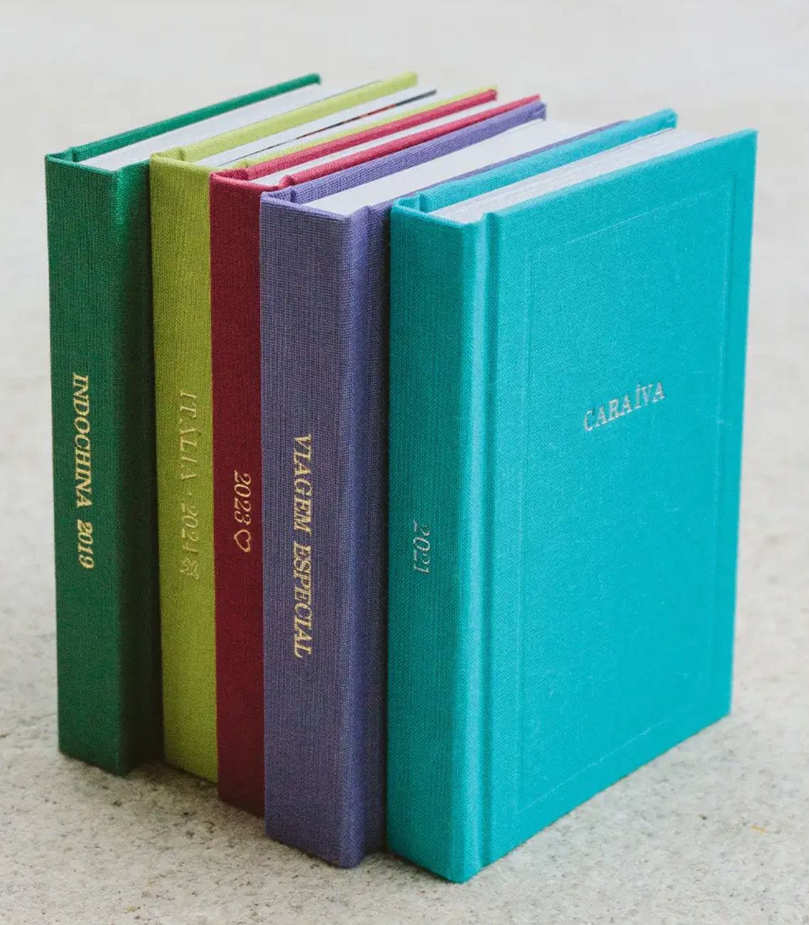 Five colorful hardcover travel journals in teal, purple, red, green, and blue, standing upright on a light surface.