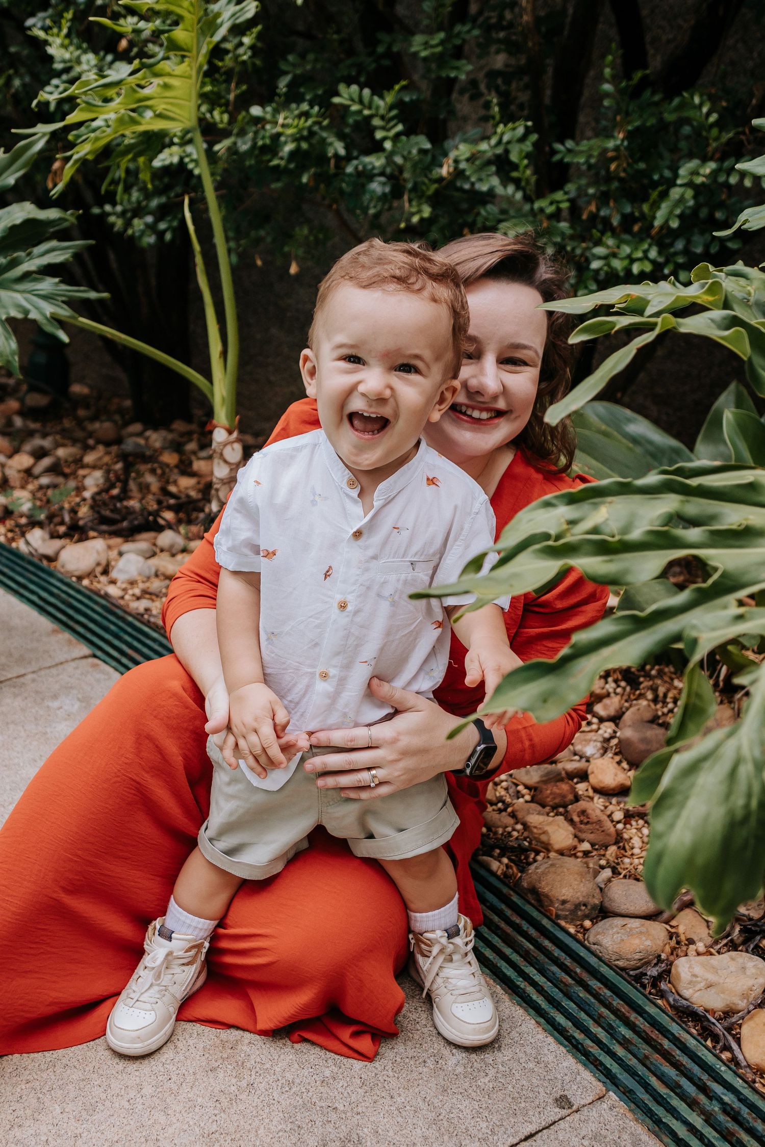 Candid outdoor photo of child laughing with joy while sitting on lap among tropical plants.
