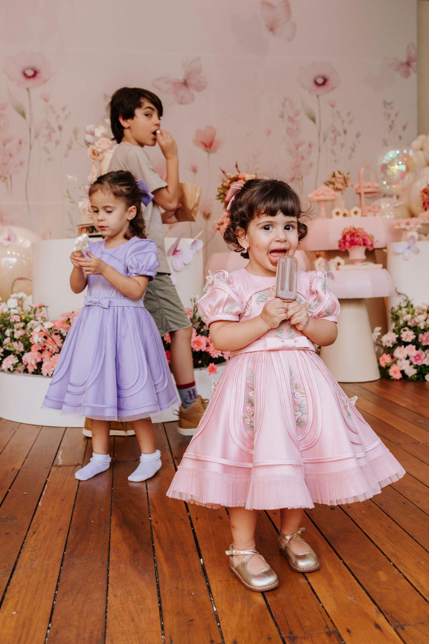 Children in pink and purple party dresses enjoying celebration with floral decorations.