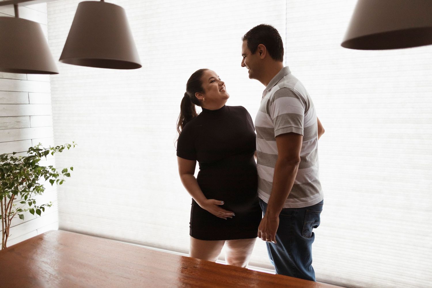 Two people stand close together near a window with hanging pendant lamps in a bright white room.