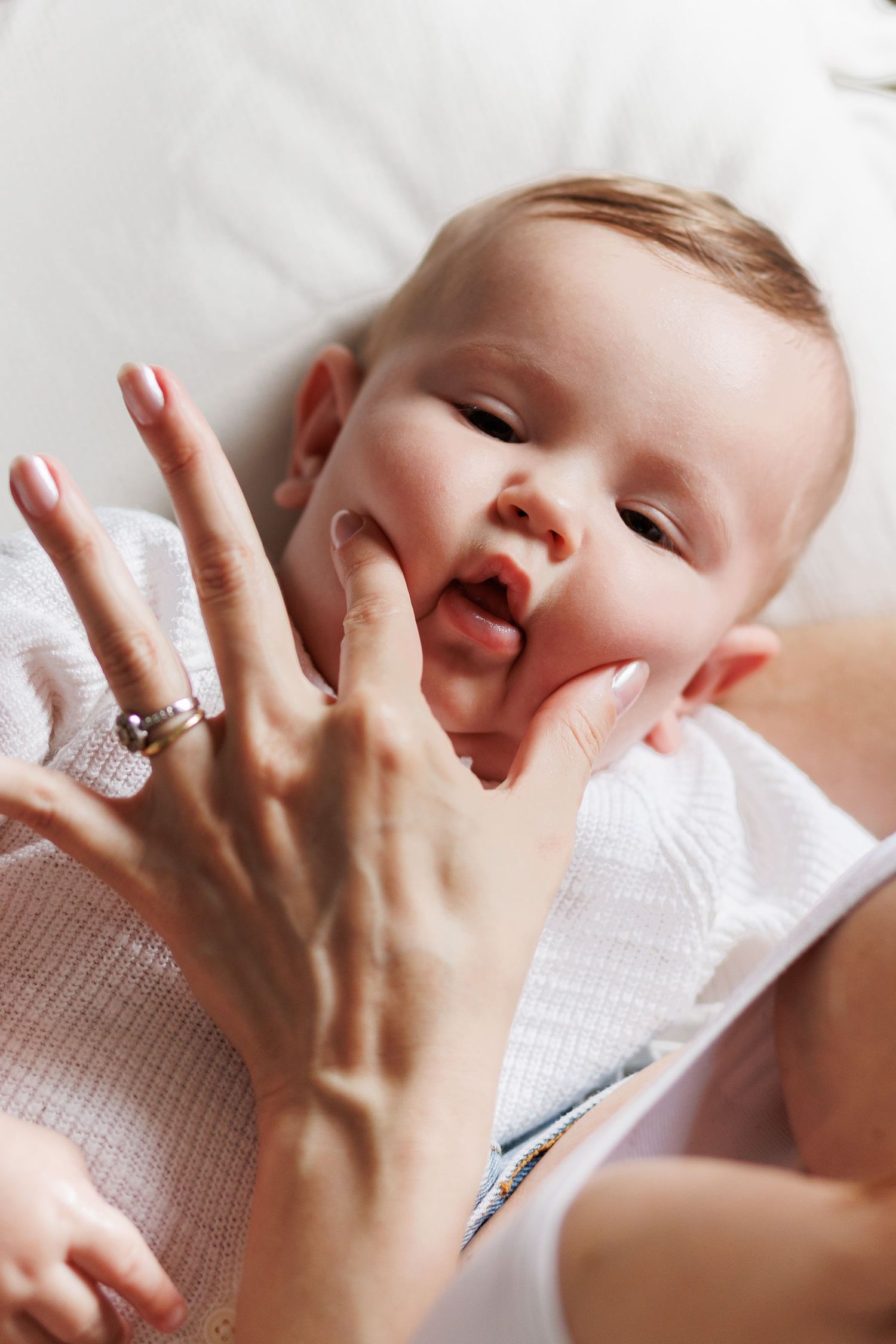 A baby lying on a white surface, playfully mouthing an adult's hand with a ring on their finger.
