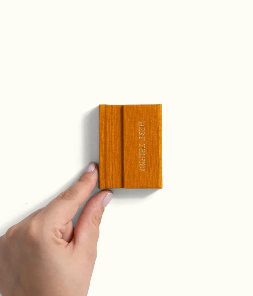 Hand holding a small mustard yellow notebook with elastic band closure and embossed text on white background.