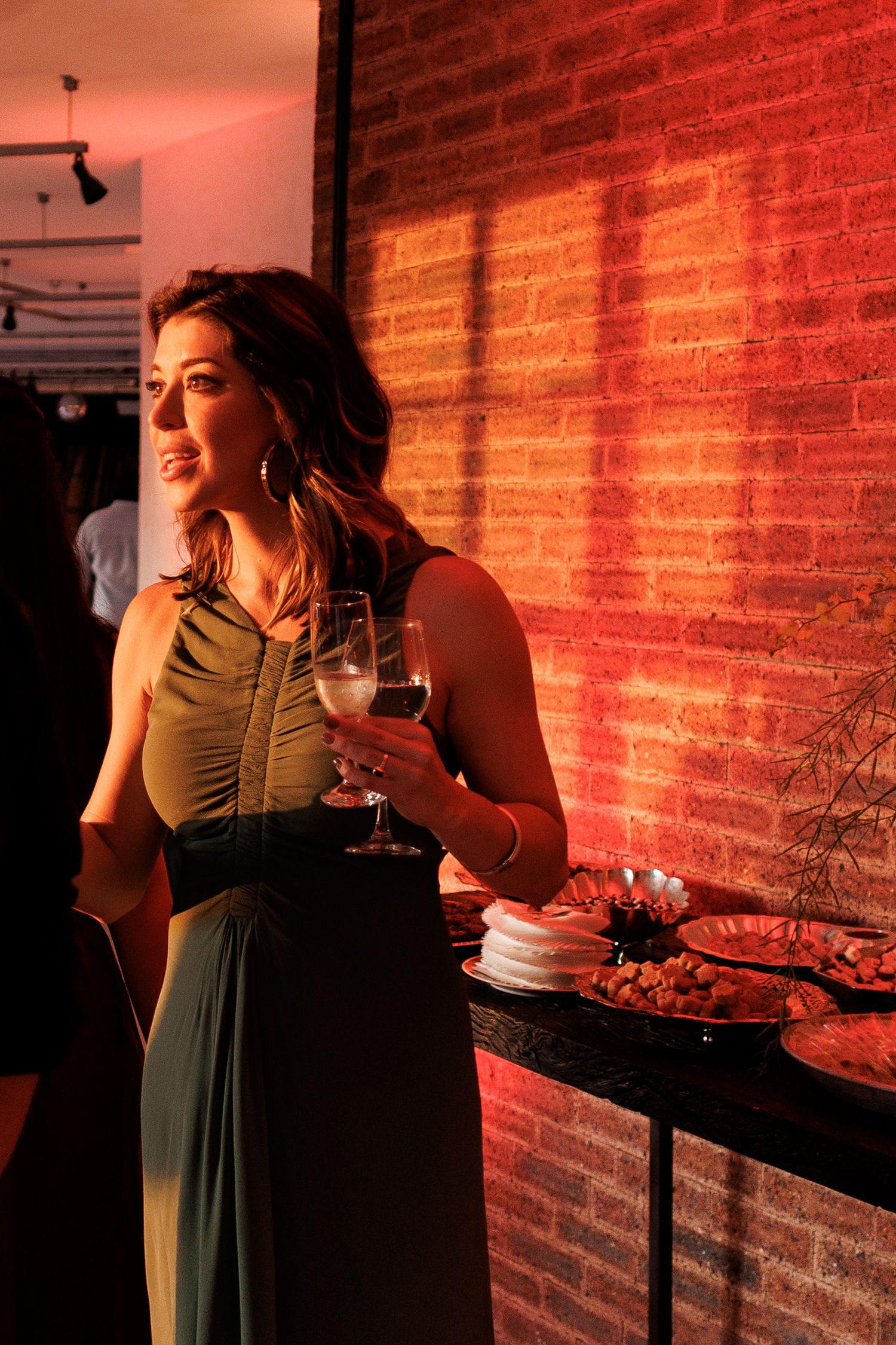 A person in a green dress holds a wine glass against a red-lit brick wall in a moody setting.
