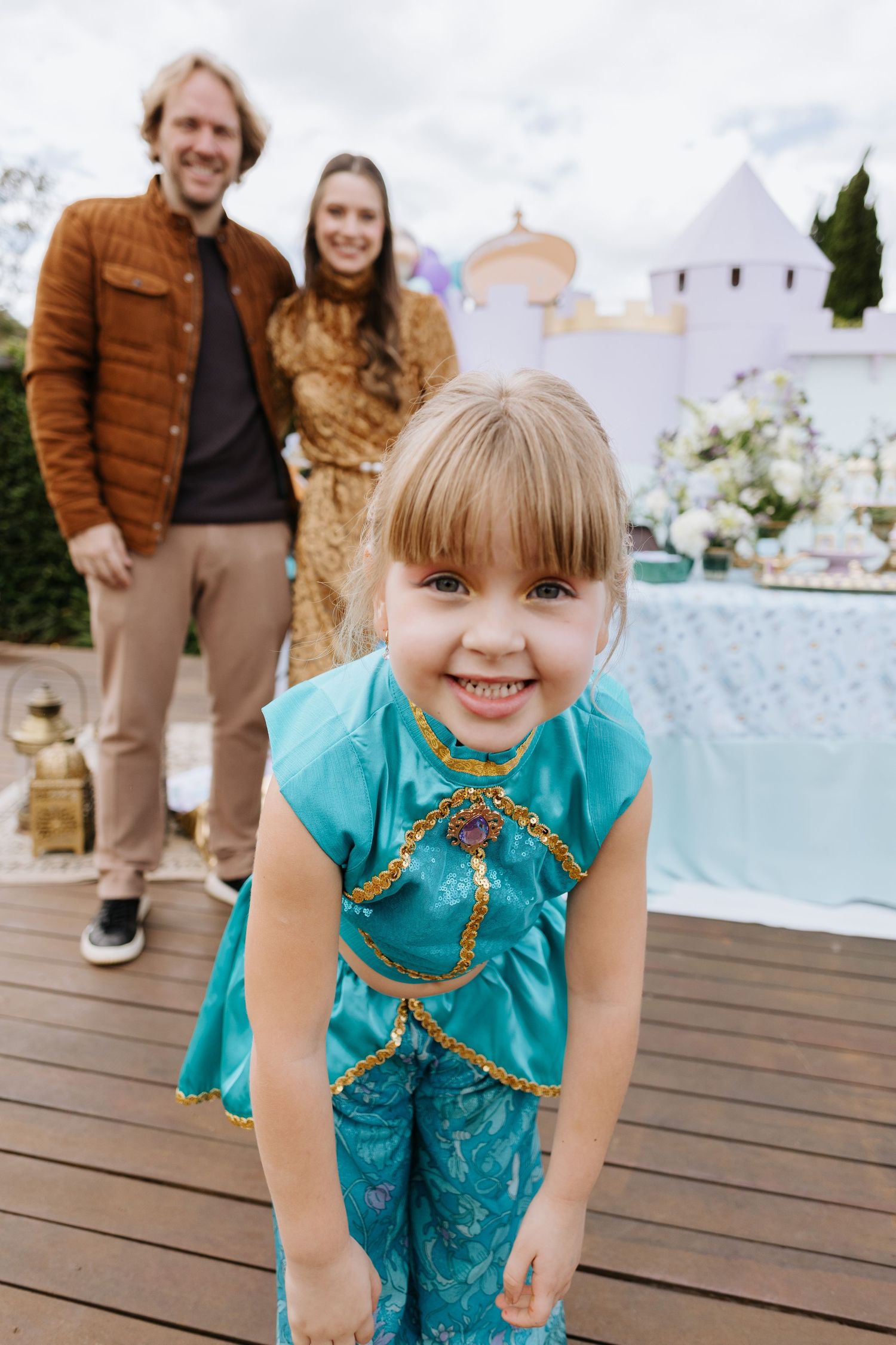 A child in a turquoise outfit plays and poses on a wooden deck near a waterfront.
