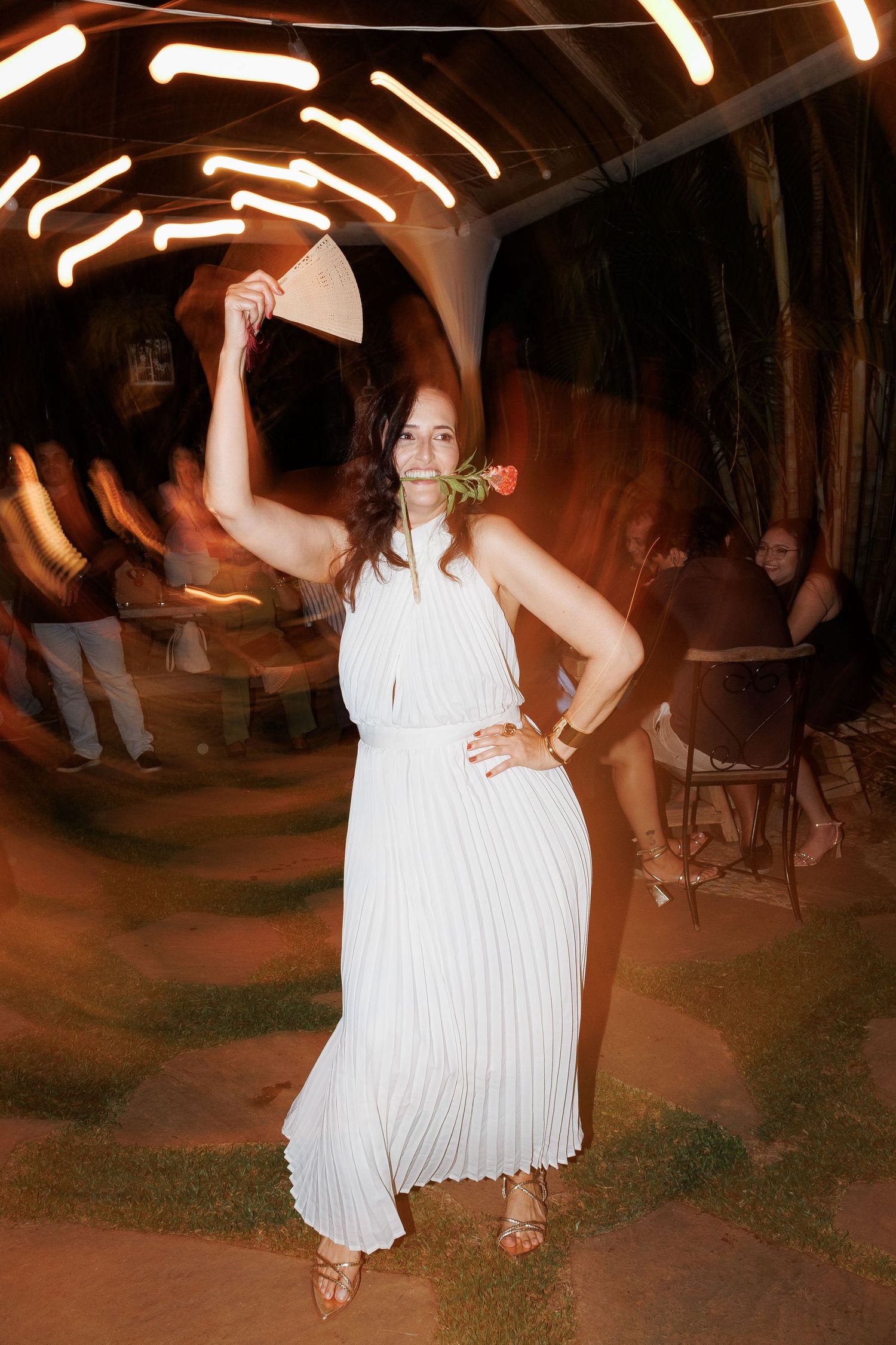 Woman in white pleated dress holding a hat, posing at an outdoor evening event with glowing lights.