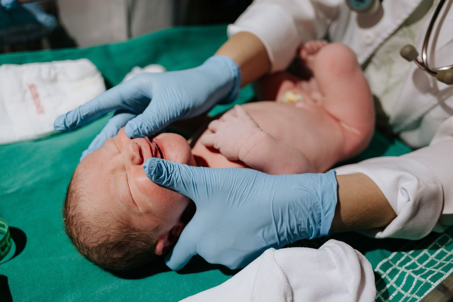 Medical professional in blue gloves examining a newborn baby on a green surgical table after delivery.