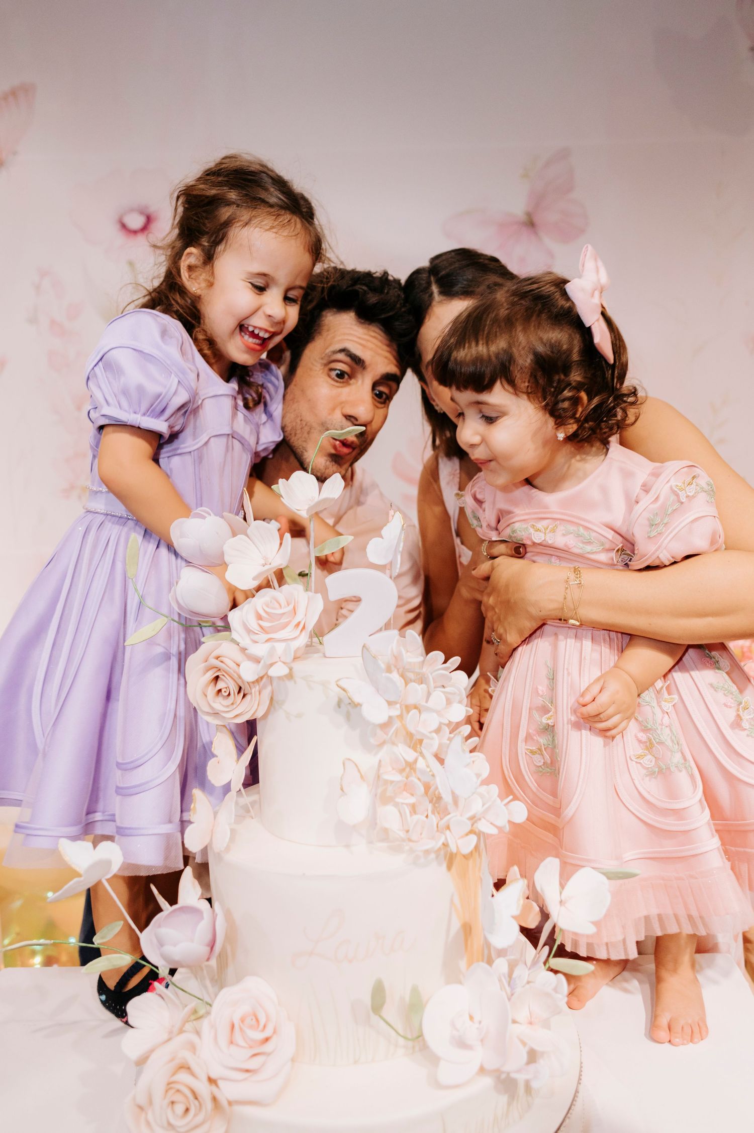 Two children in fancy dresses join an adult in playfully blowing on a white wedding cake decorated with roses.