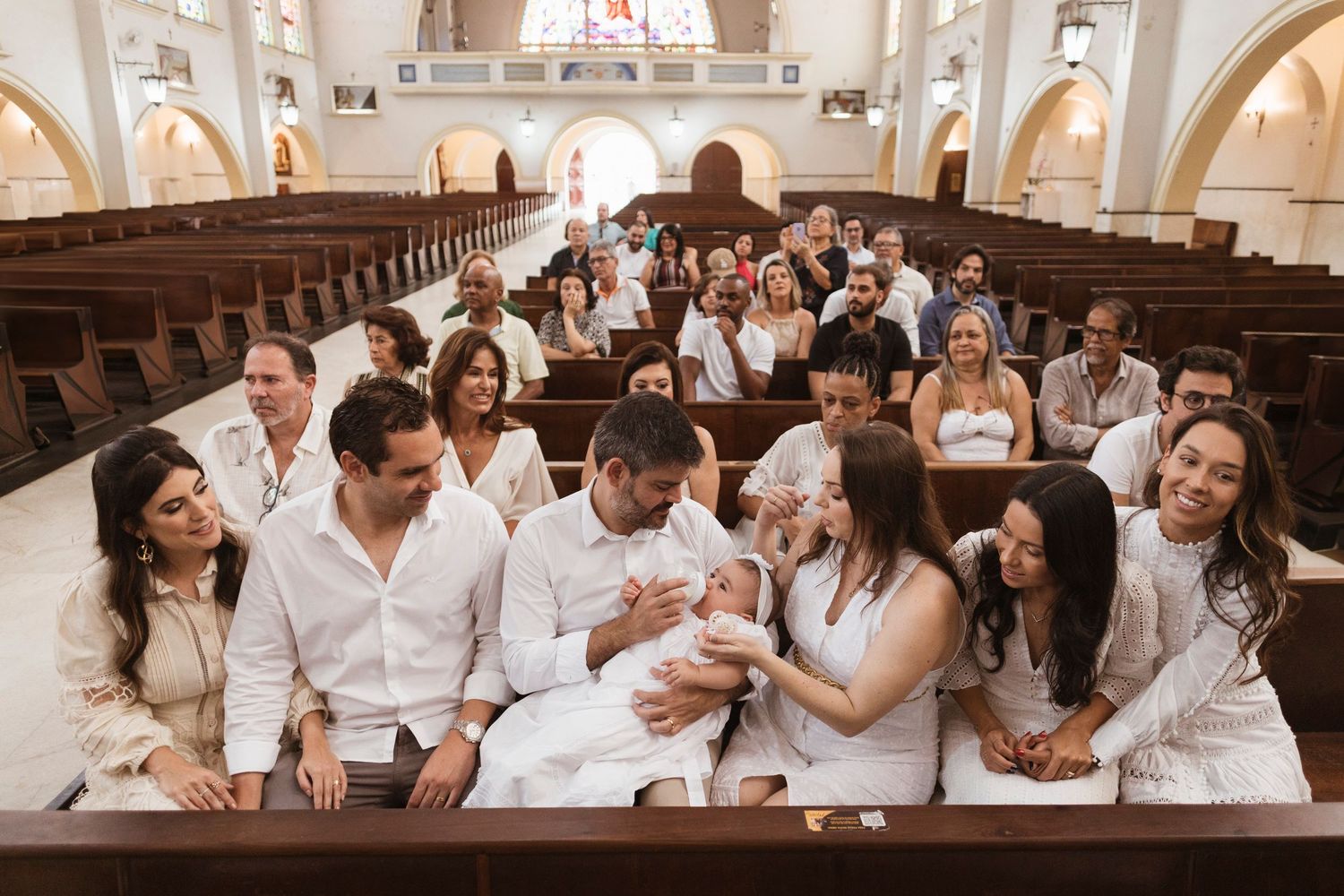 Large group in white attire gathers in church pews for baptism celebration.