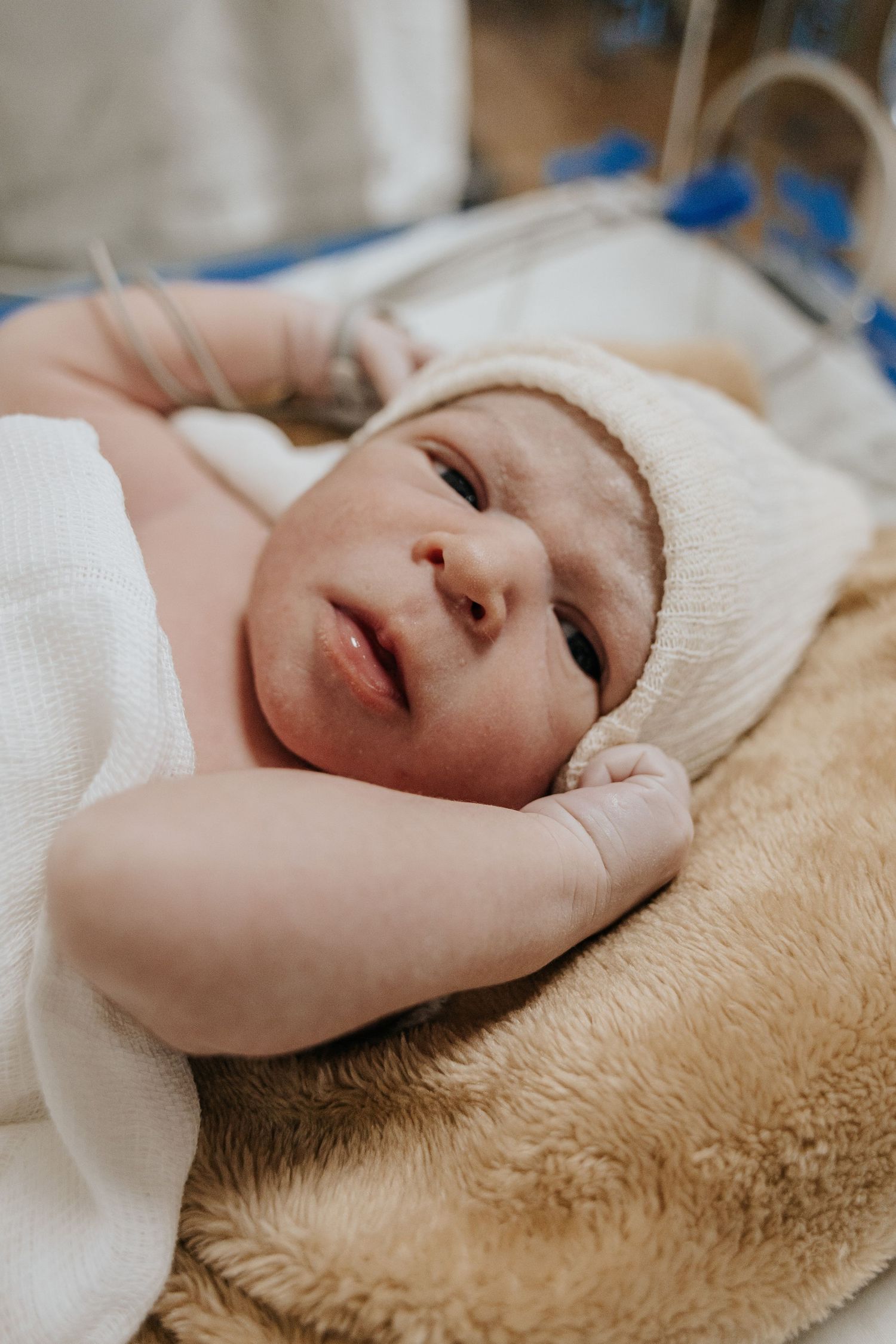 Close-up of alert newborn baby wearing white cap while lying on soft hospital bedding.