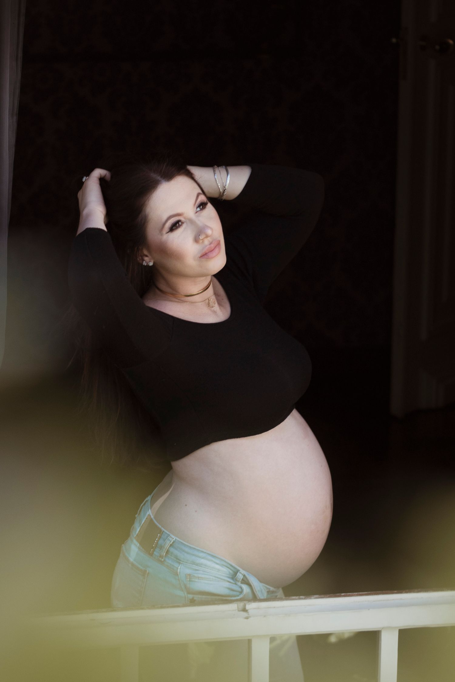 Maternity photo of pregnant woman in black top leaning against railing in moody lighting.