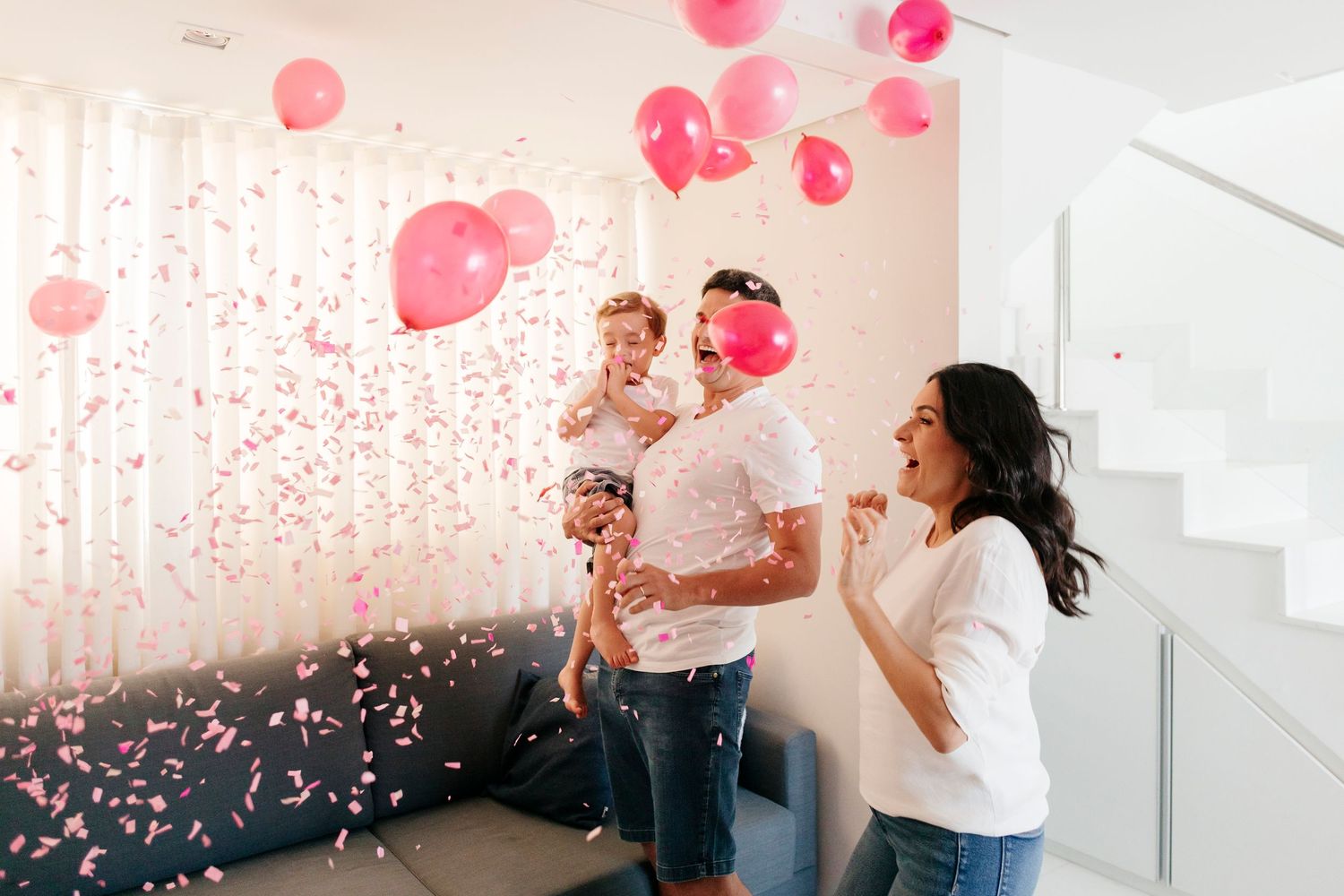Happy couple celebrates with pink confetti and balloons in a bright white modern space.