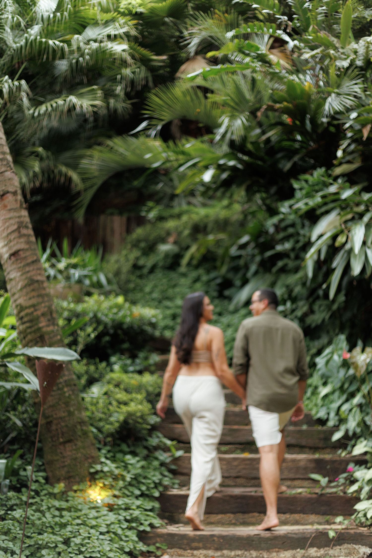 People walking along a lush tropical garden path surrounded by palm trees and dense foliage in natural lighting.