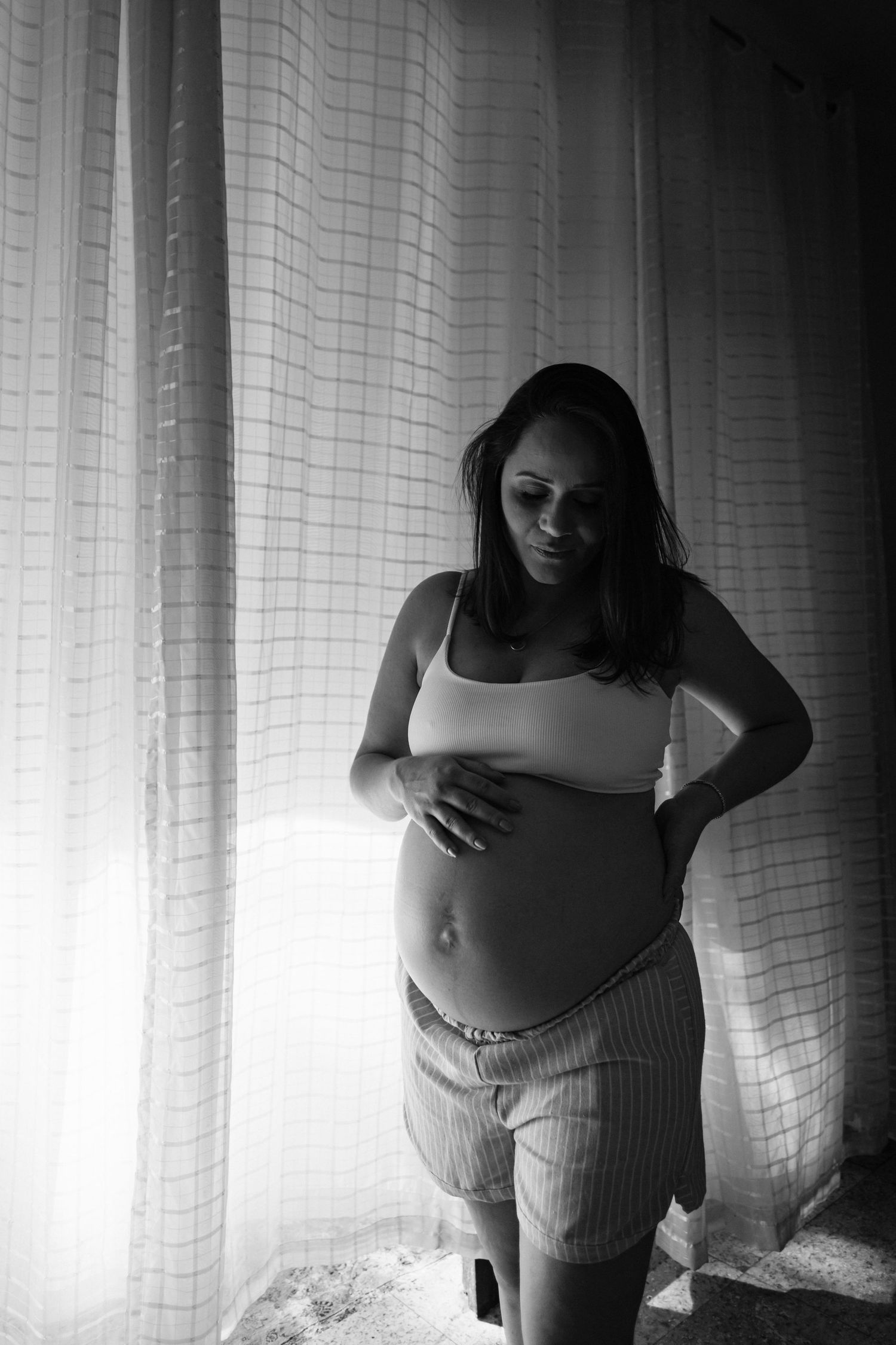 Monochrome portrait of expectant mother in casual wear standing by window with natural lighting.