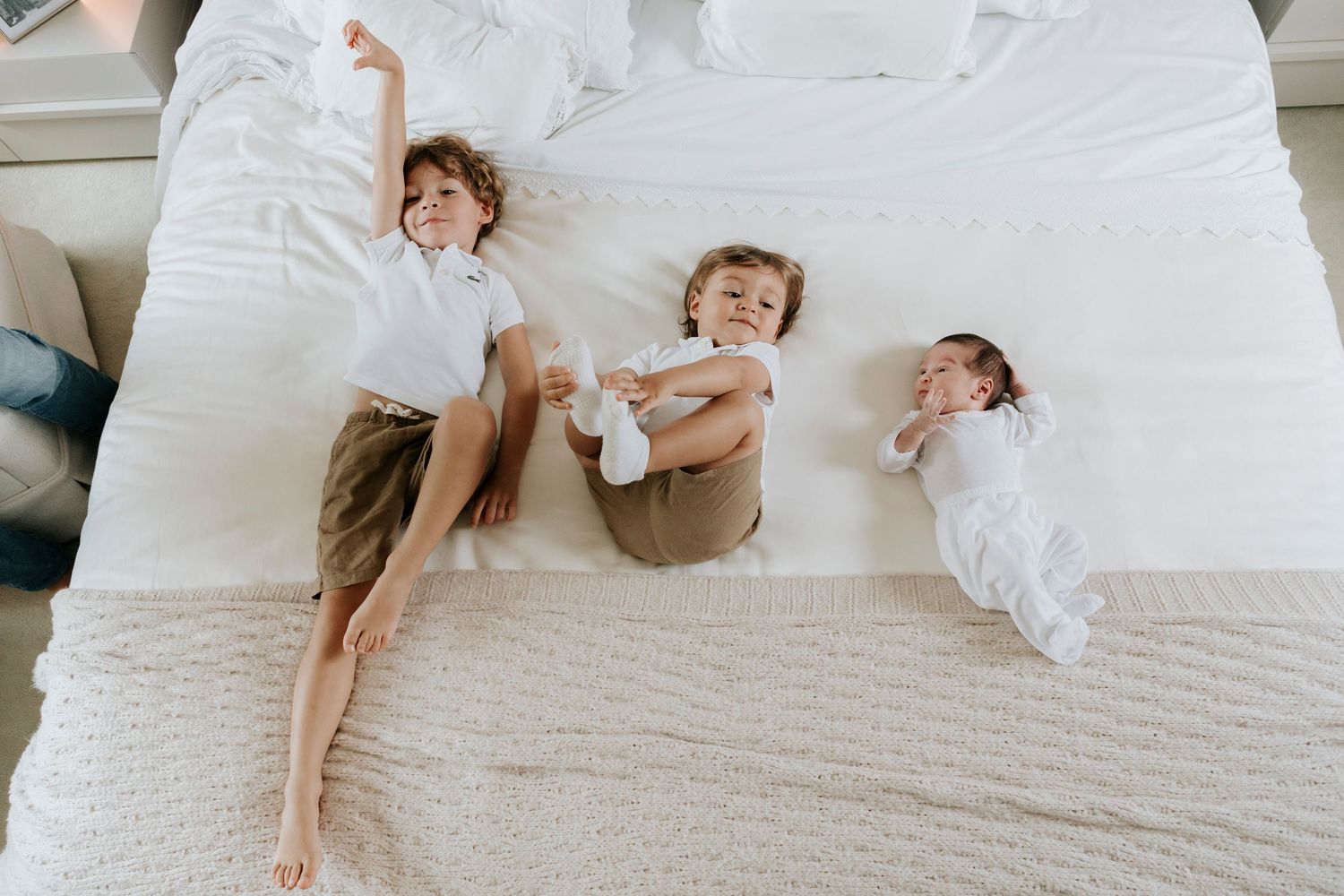 Three children lying playfully on white bed while laughing and smiling.