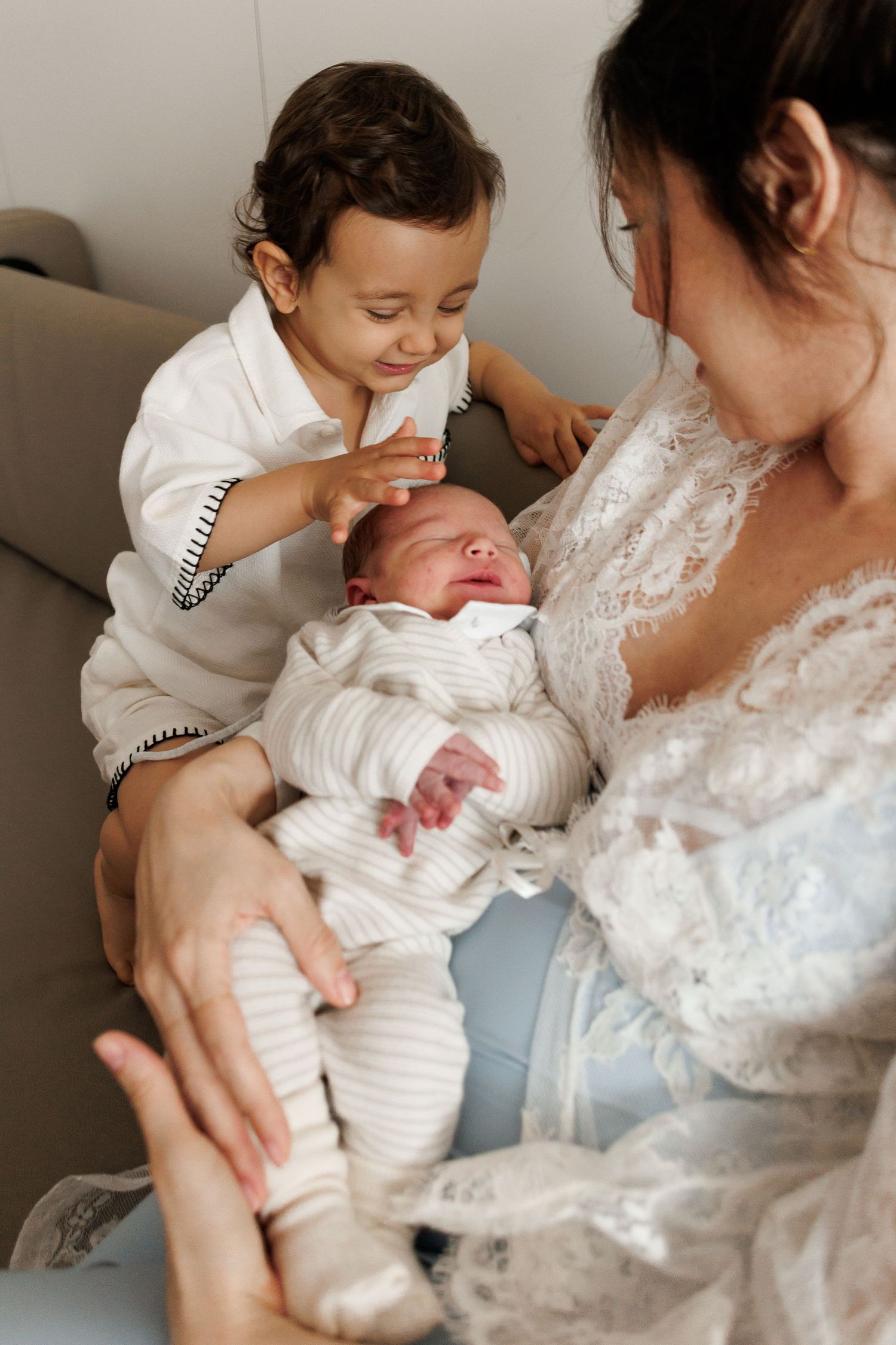 A mother in lace robe holds her newborn while a toddler boy gently touches the baby's head, smiling warmly.