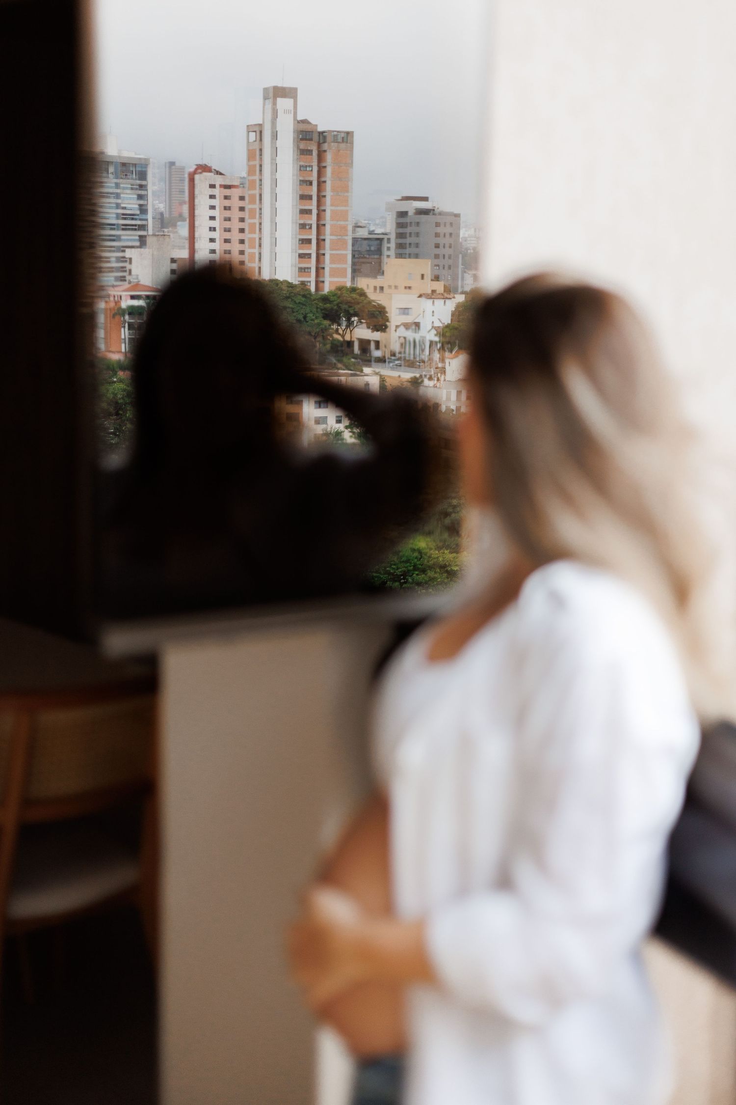 Pregnant woman in white shirt gazing at urban cityscape through apartment window.
