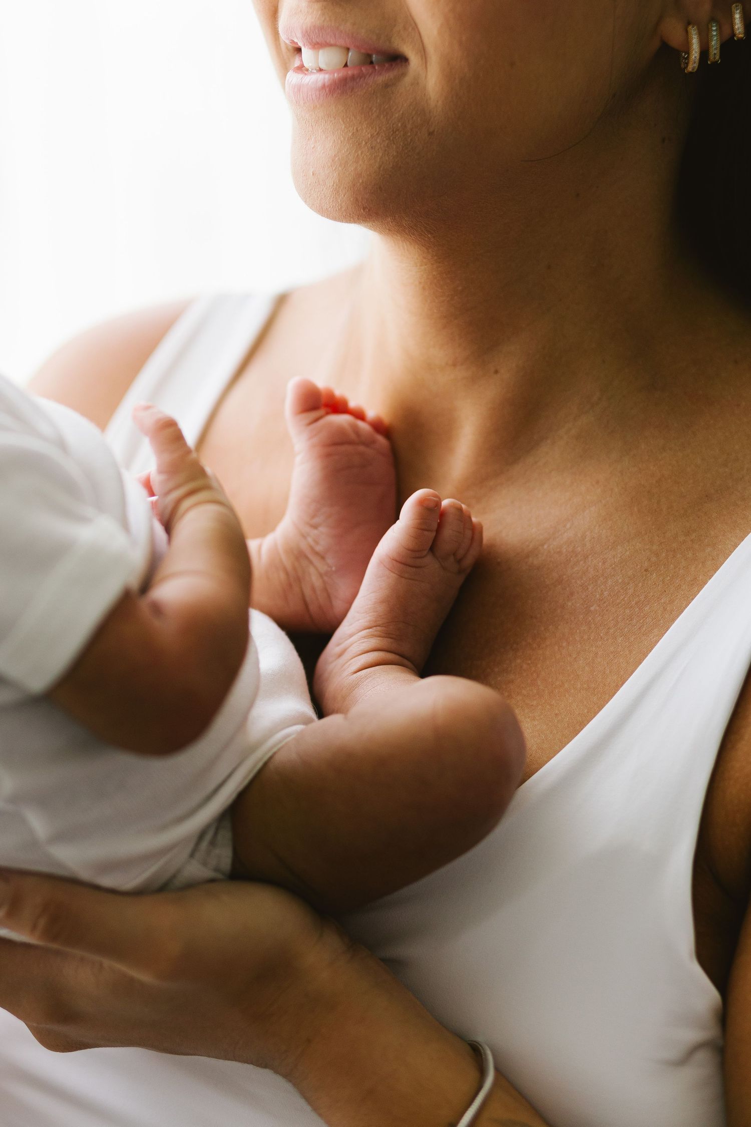 Smiling mother in white tank top cradling newborn baby, with tiny infant feet visible against her chest.