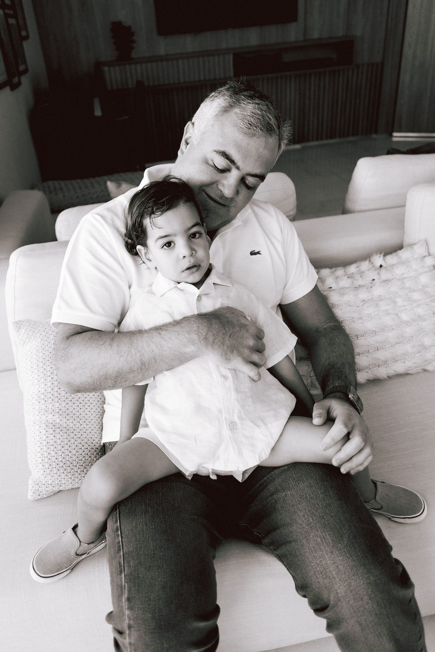 Black and white photo of a grandfather lovingly holding a young toddler on his lap in a cozy living room.