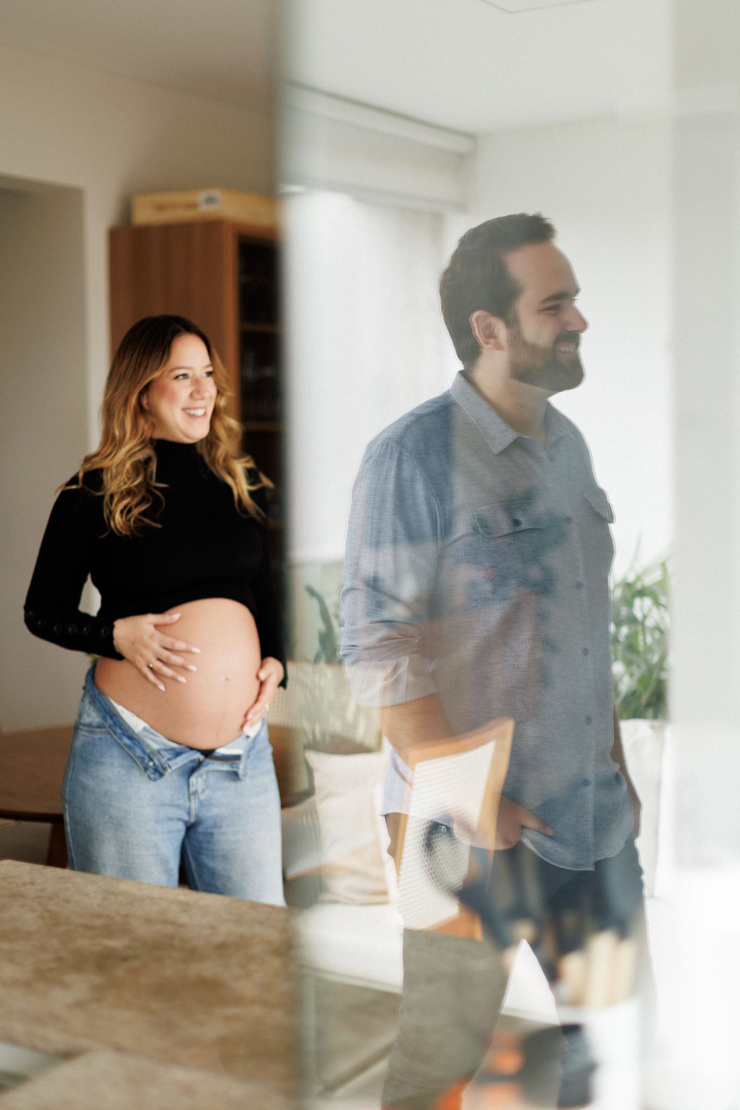 Smiling pregnant woman in black top and jeans cradles her baby bump beside a man in a denim shirt indoors.
