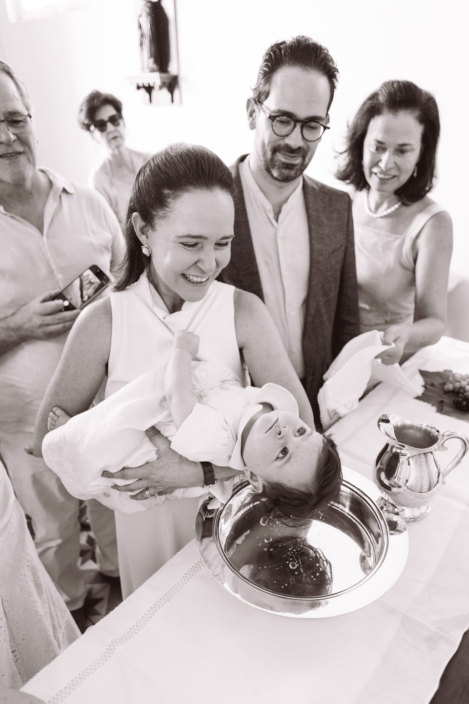 Black and white photo of a baby baptism ceremony with family gathered around a christening font in a church.