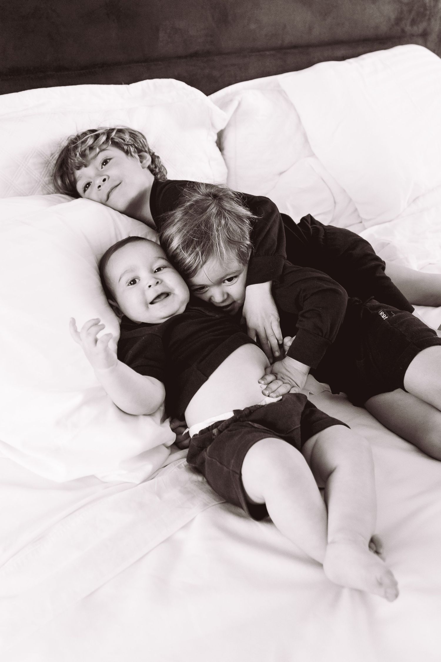 Black and white photo of three young siblings cuddling together on a white bed, sharing a sweet moment.