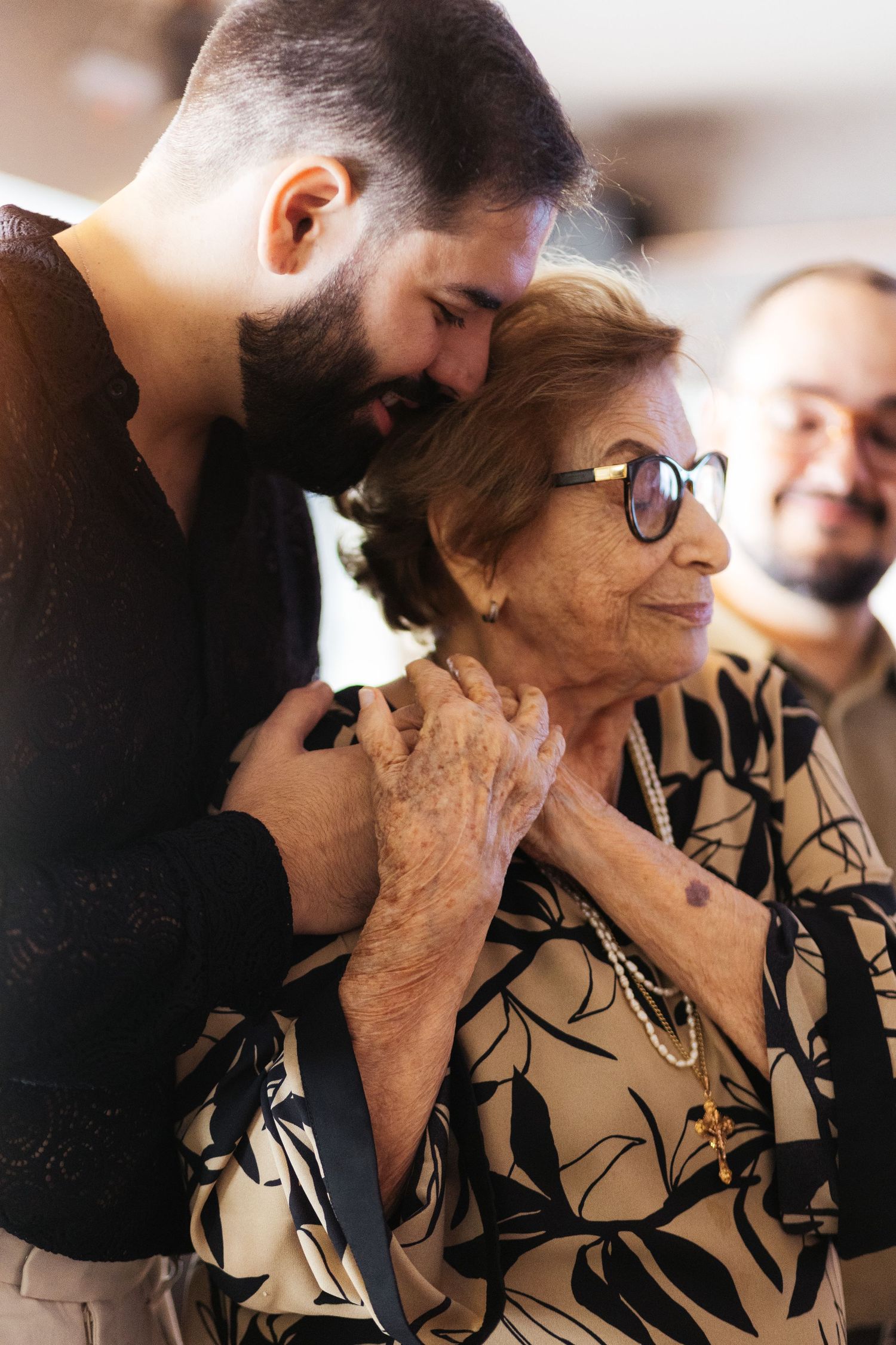 Young bearded man tenderly embracing elderly woman wearing glasses and floral blouse at family gathering.
