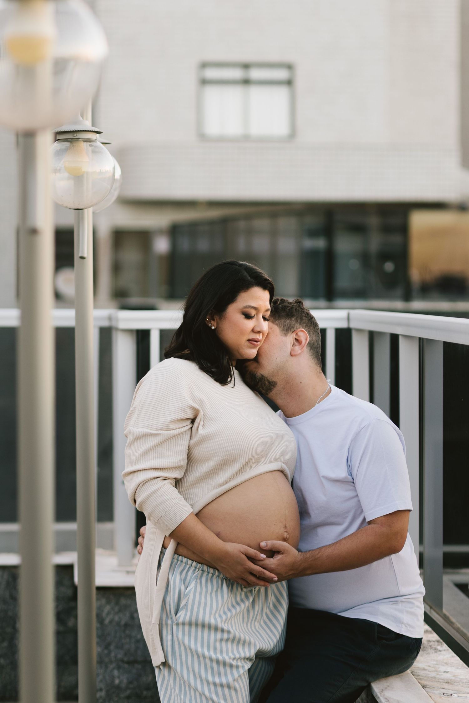 A man kisses a pregnant woman's cheek while cradling her baby bump outdoors on a balcony.