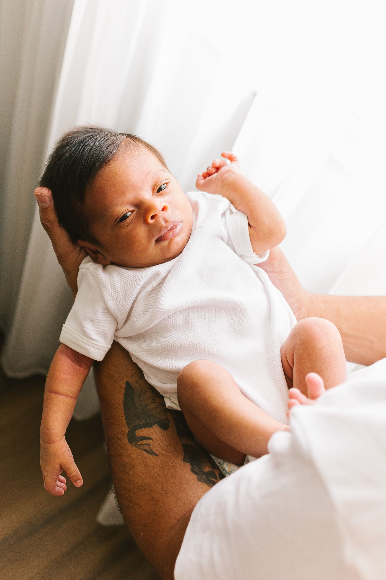 Newborn baby in white onesie resting on parent's tattooed arm near bright window.