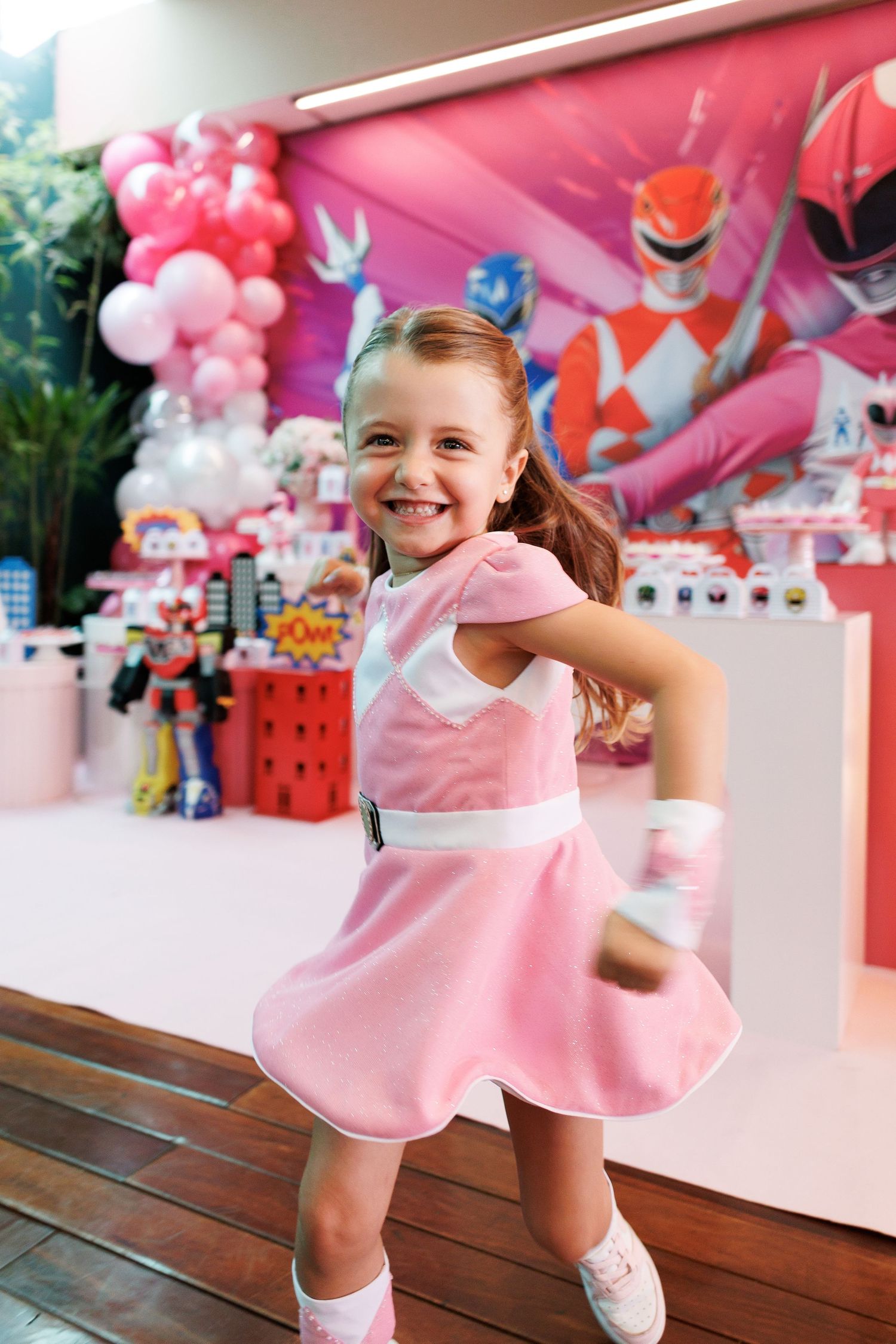 Young girl in pink Power Ranger costume smiling at birthday party with themed decorations and balloon backdrop.