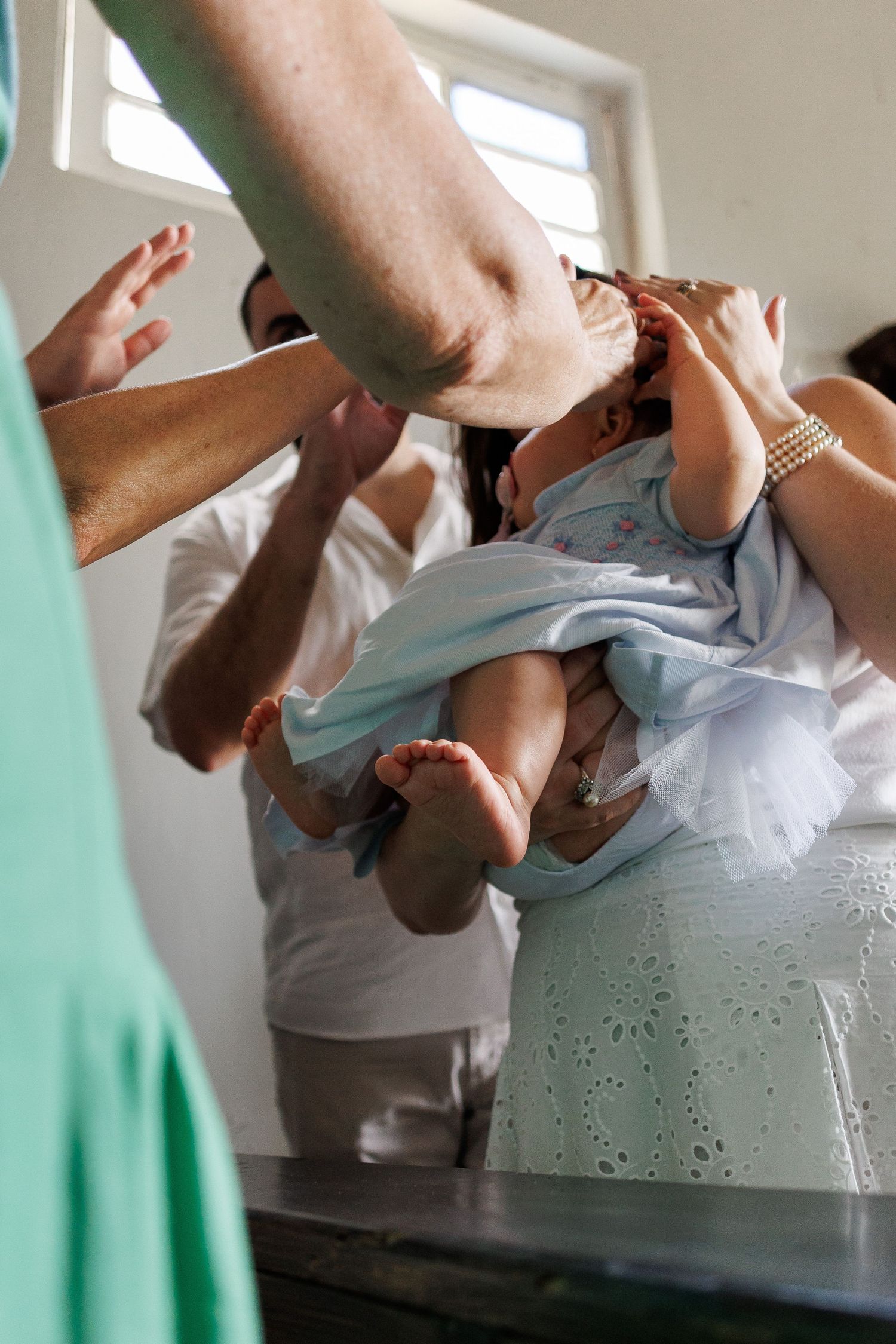 A baby in a white dress being held during a baptism ceremony with adults surrounding them.
