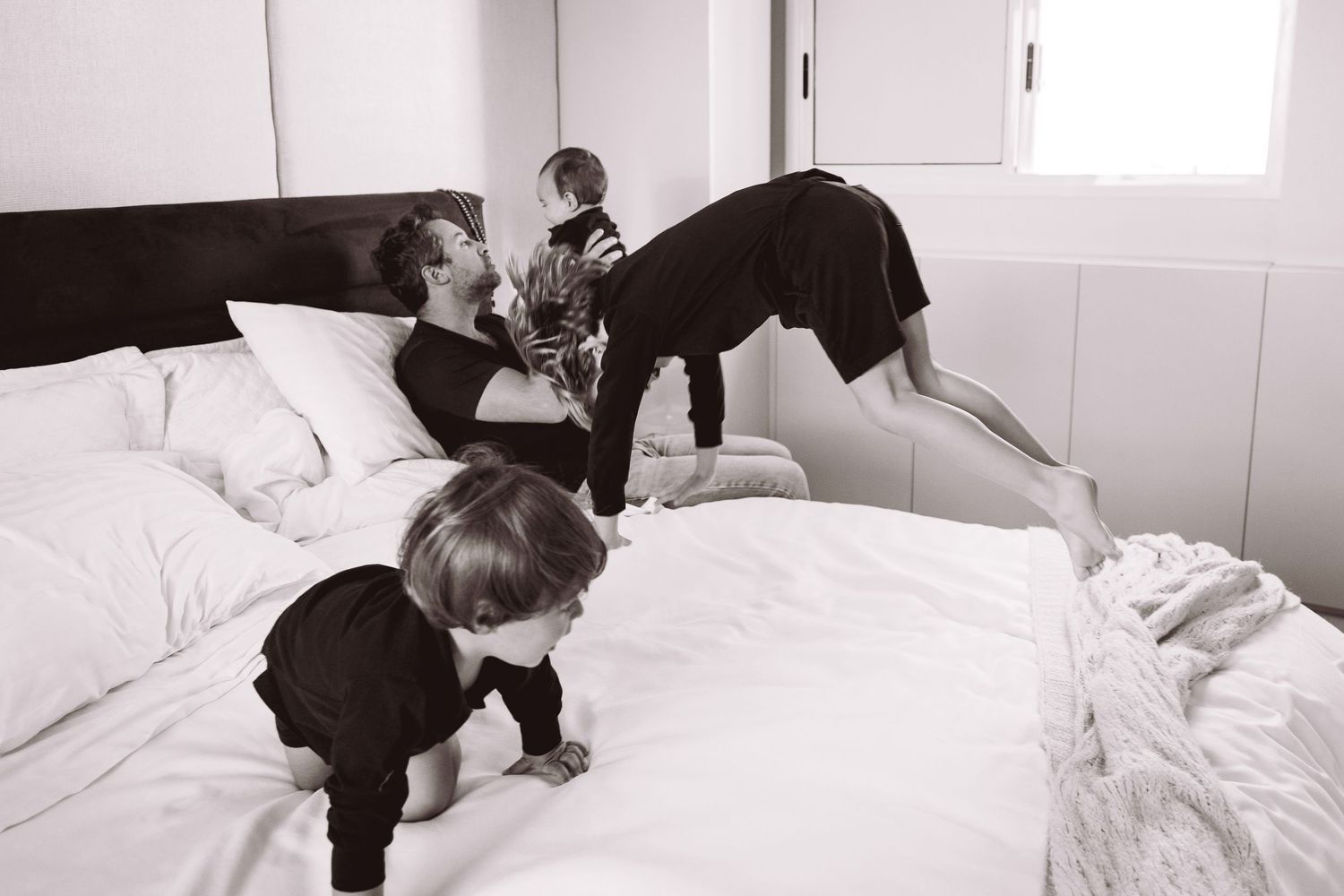 Black and white photo of a father playfully wrestling with three young children on a white bed.