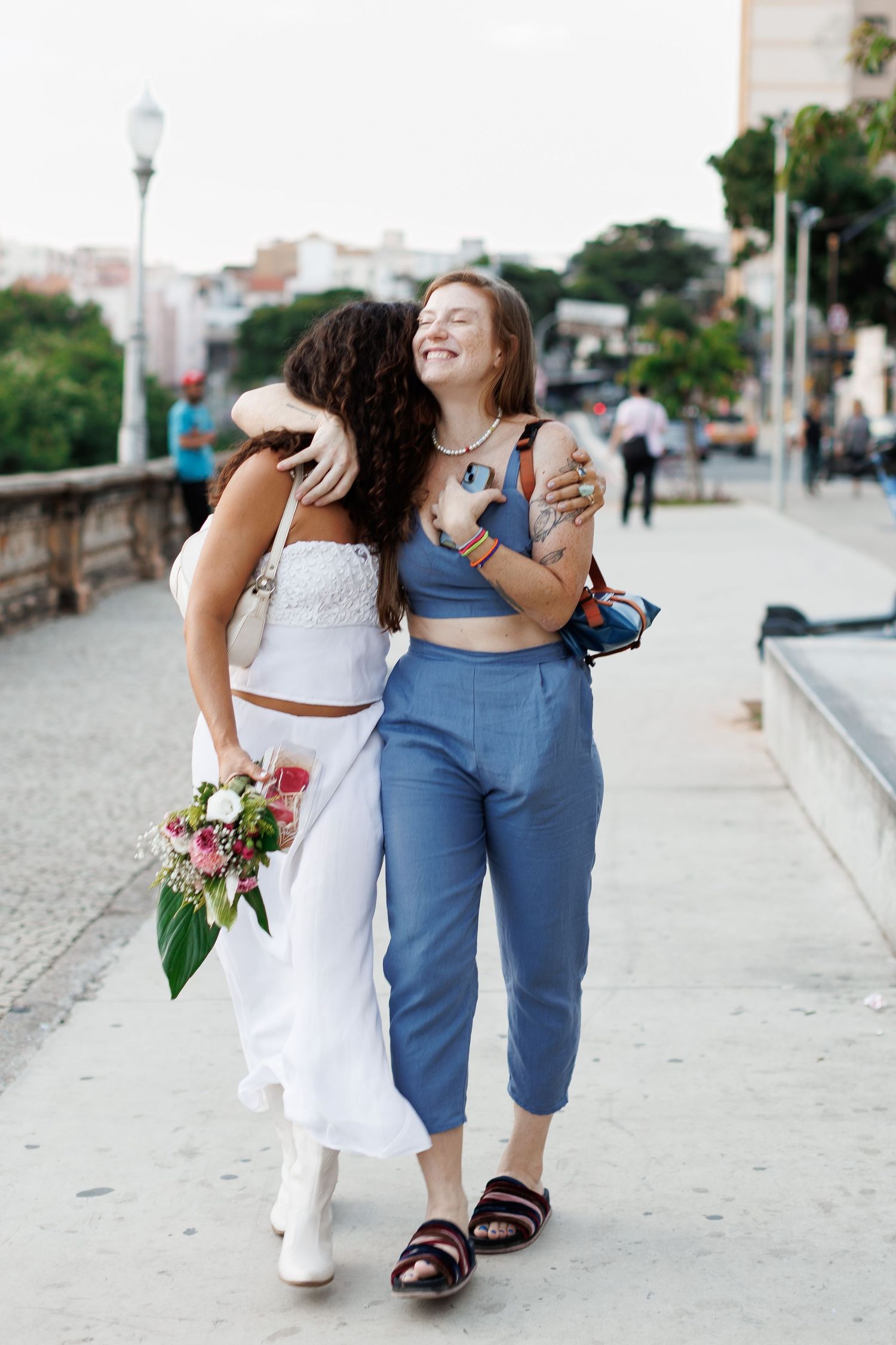 Two women embracing and walking together outdoors, one in white dress holding flowers, other in blue crop top and pants.
