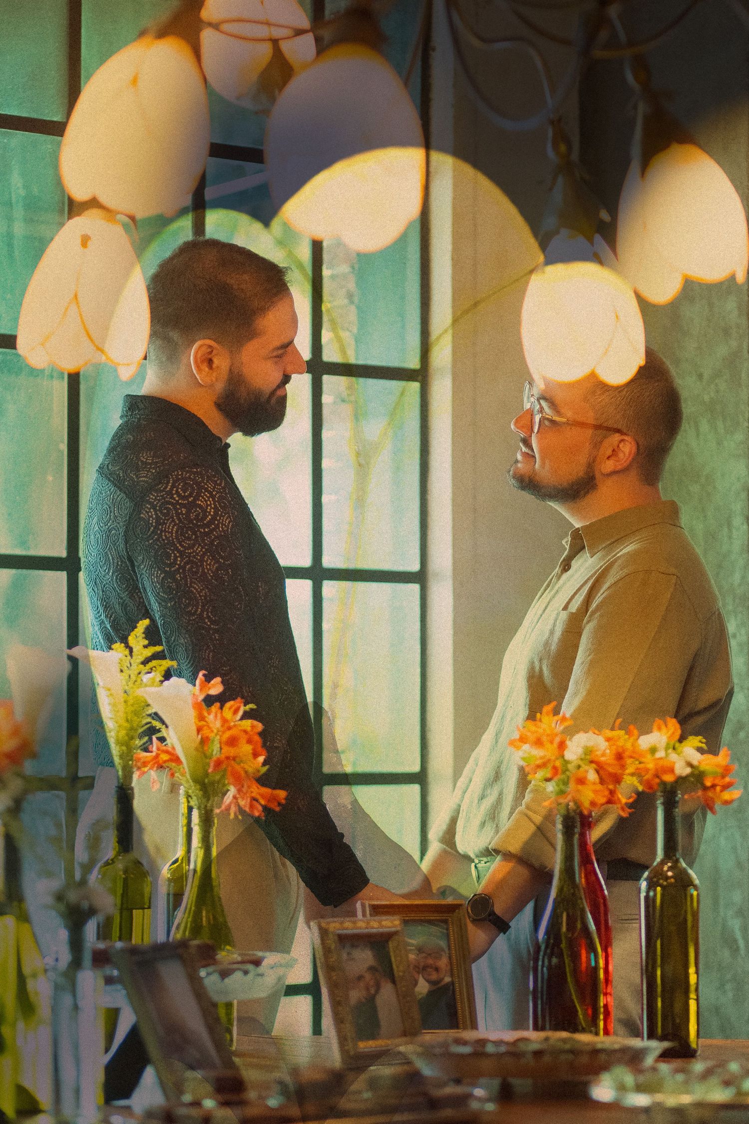 Two men holding hands and facing each other surrounded by orange flowers and warm pendant lights in an intimate setting.